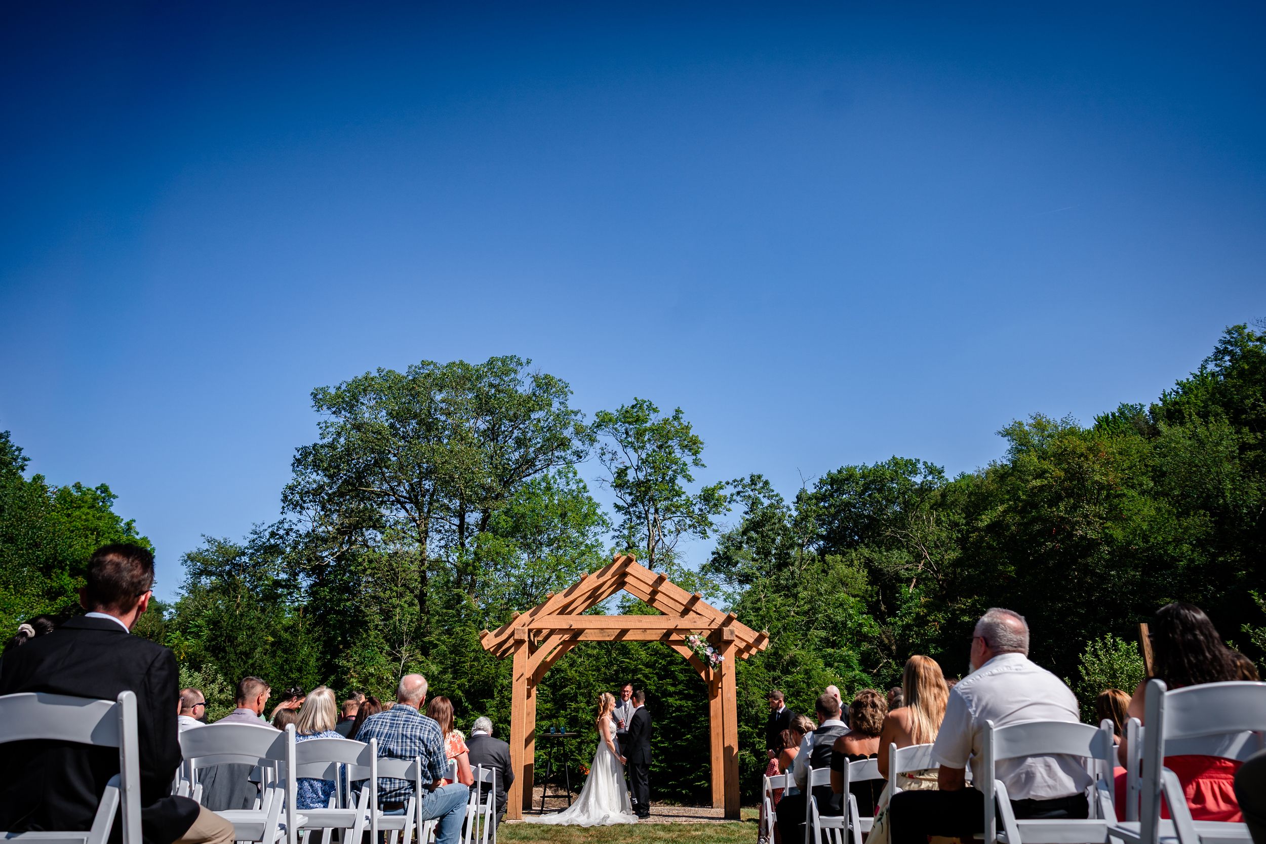 Panoramic view of an outdoor wedding ceremony setup with wooden arch and white chairs.