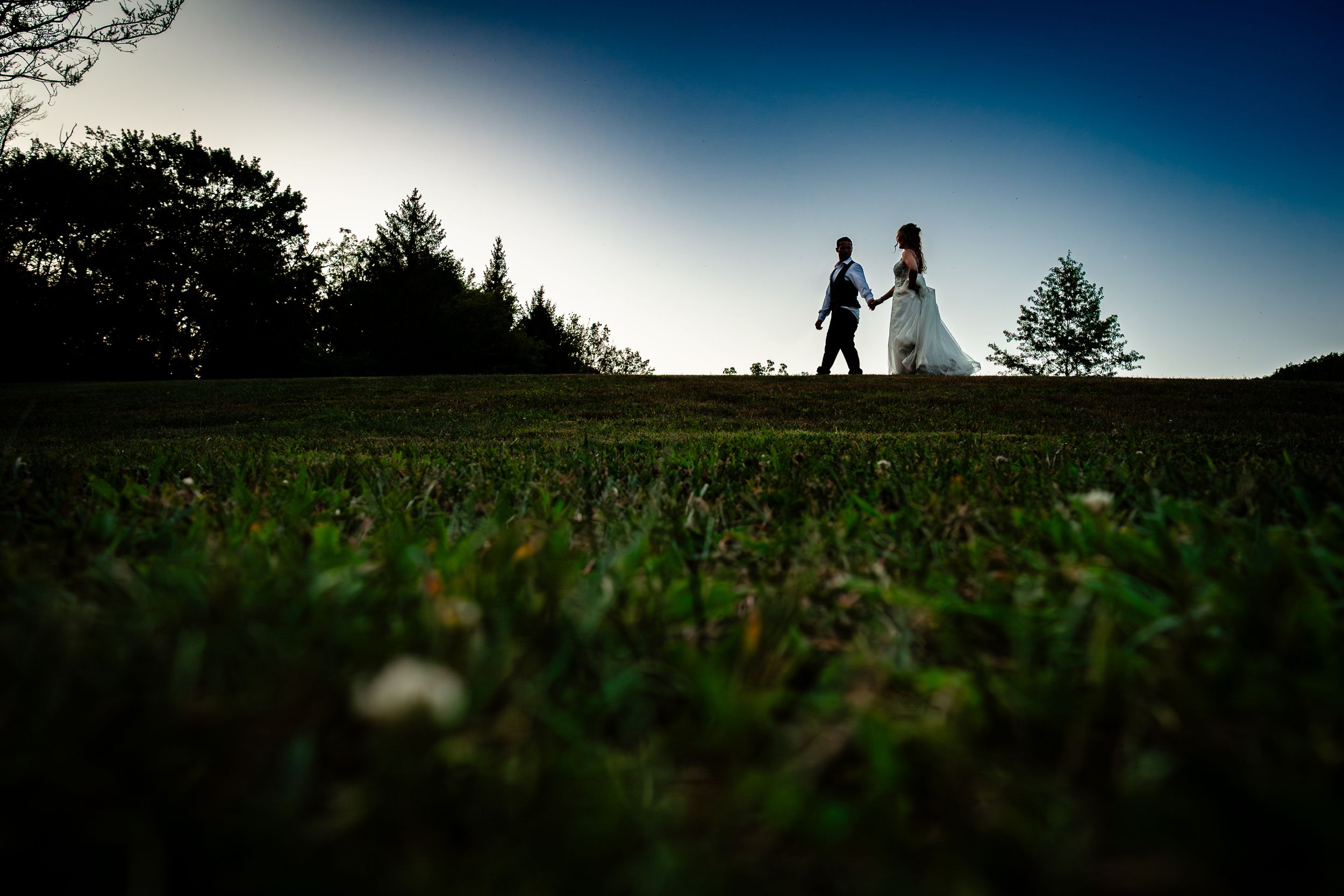 Silhouettes of a wedding couple stand against a dramatic sunset sky in an outdoor setting.