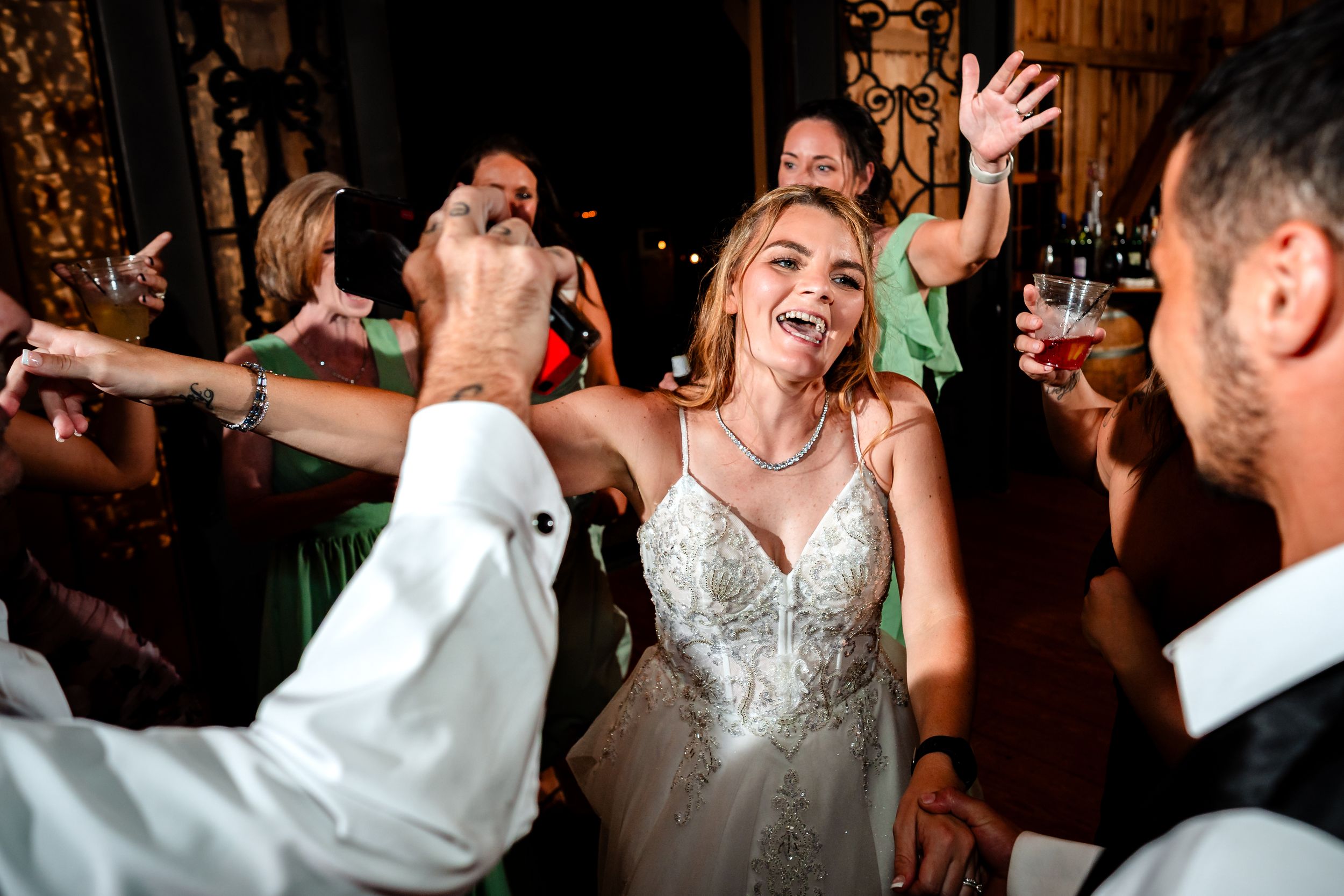 Wedding party celebrates with energetic dancing under string lights in rustic barn venue.