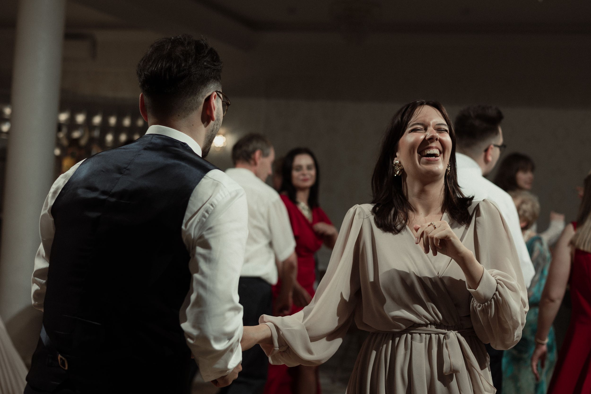 A candid moment at a wedding reception shows a guest in a white dress laughing while dancing with others on the dance floor.