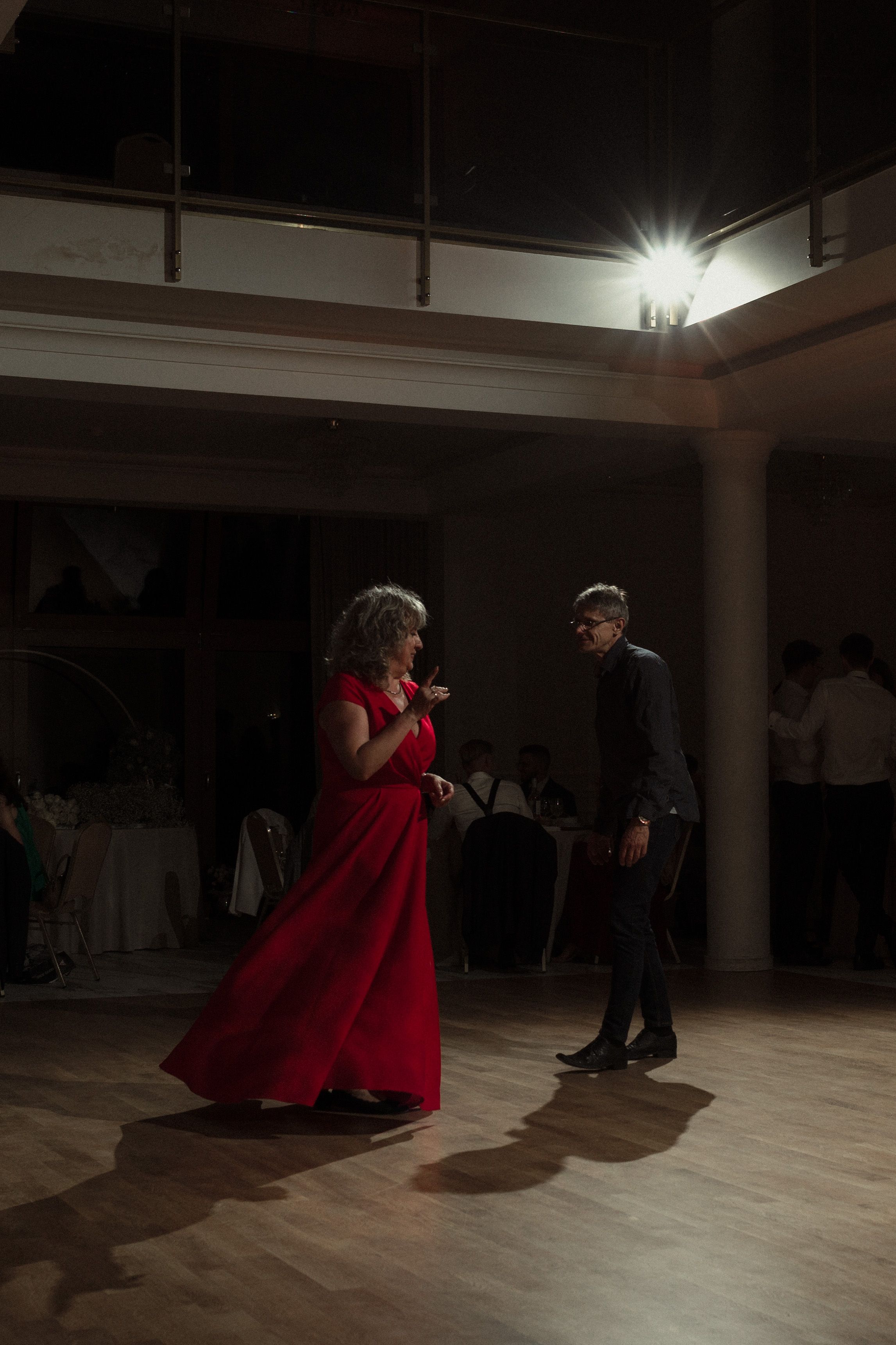 Dancers move across a wooden ballroom floor as one performer in a flowing red dress takes the spotlight.