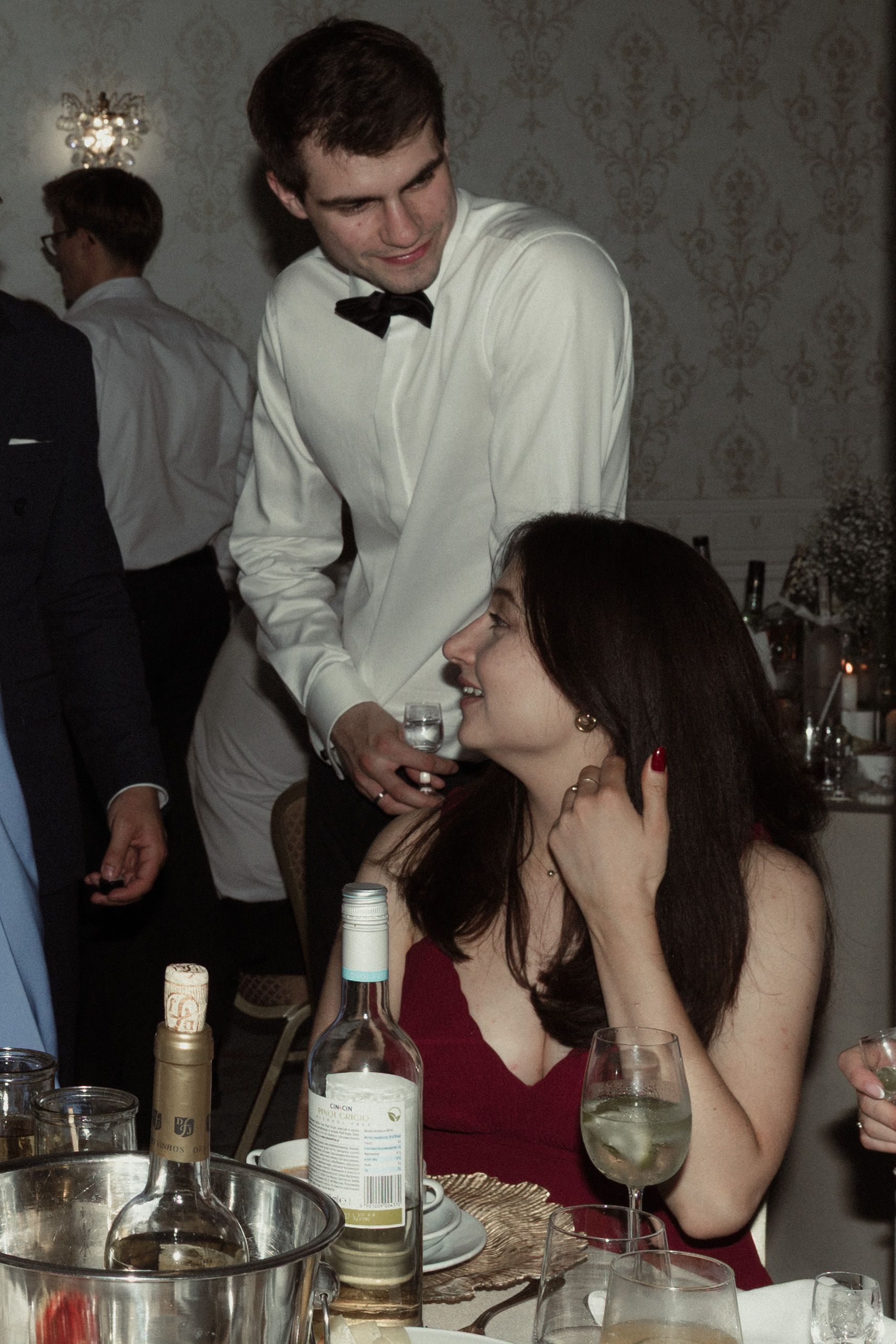 A server in a white dinner jacket tends to diners at an elegant evening event with wine bottles on the table.