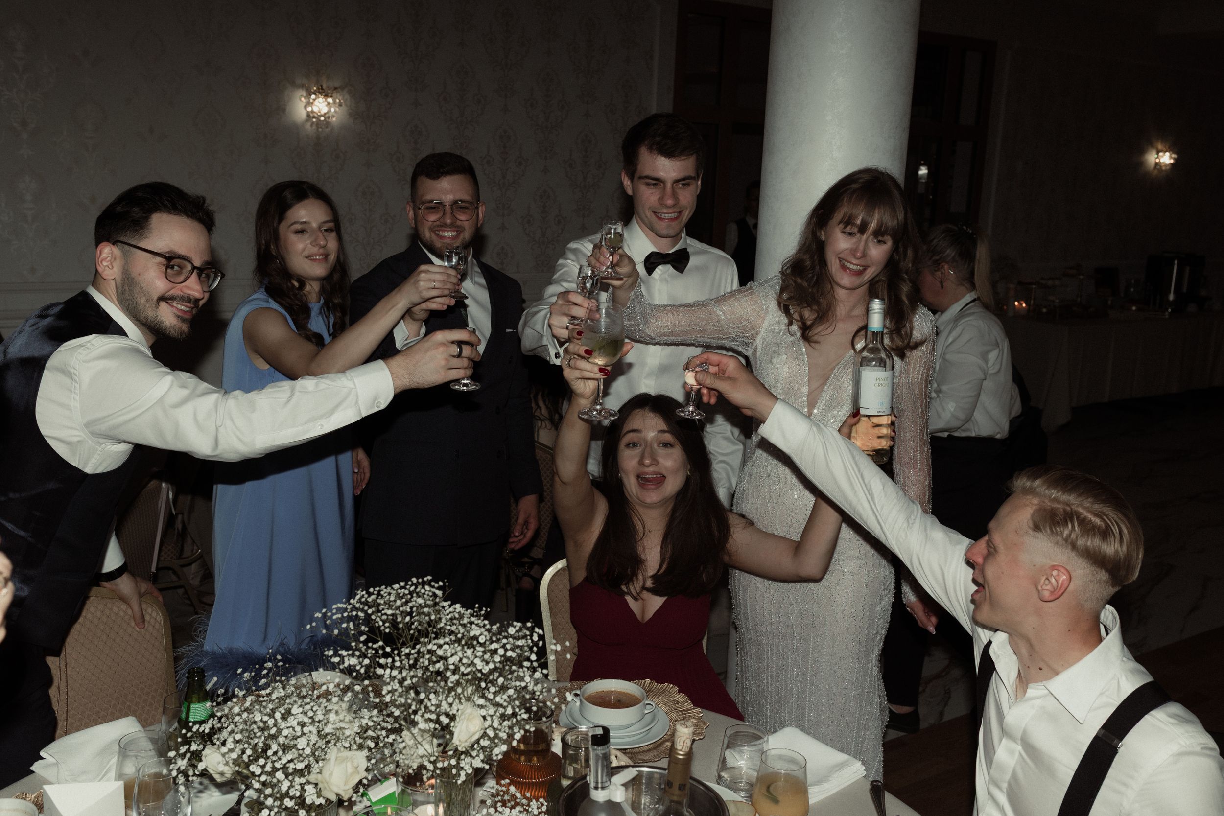 A group of people dancing and celebrating at a dimly lit party with white lights visible in the background.
