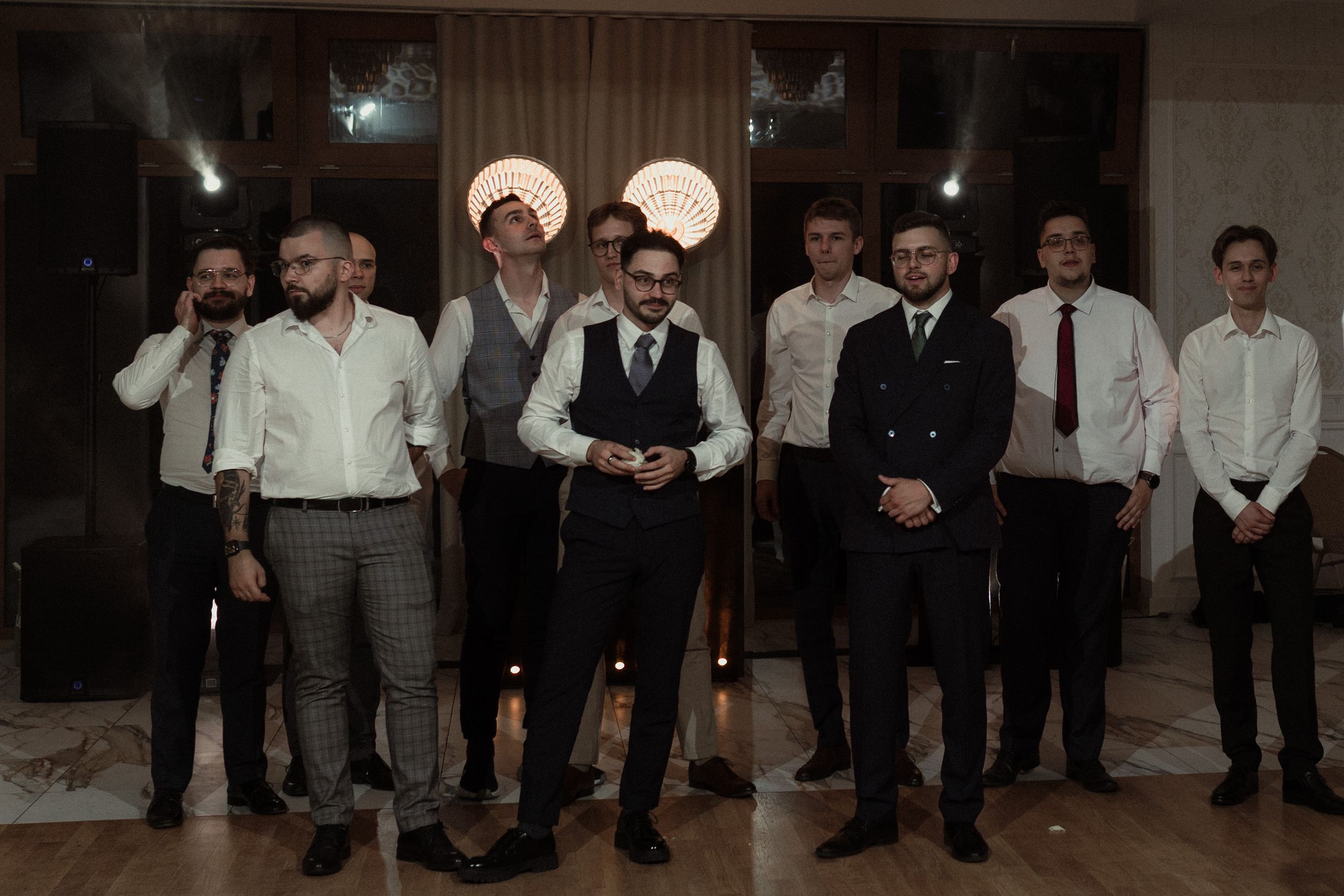 A wedding party of groomsmen in formal attire stand together in a dimly lit indoor venue.