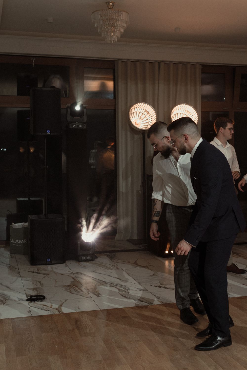 People gather on a dance floor with decorative spherical lights and a chandelier hanging overhead at an elegant evening event.