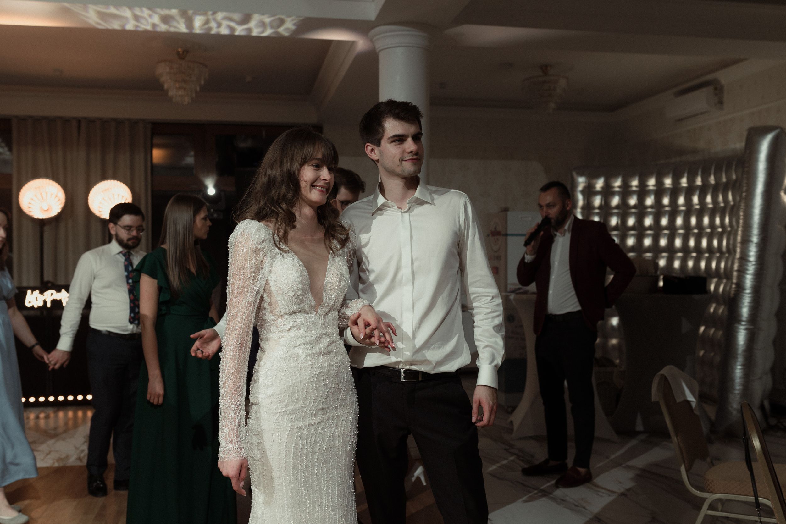 A couple dancing together at an indoor reception venue with decorative lighting and guests in the background.