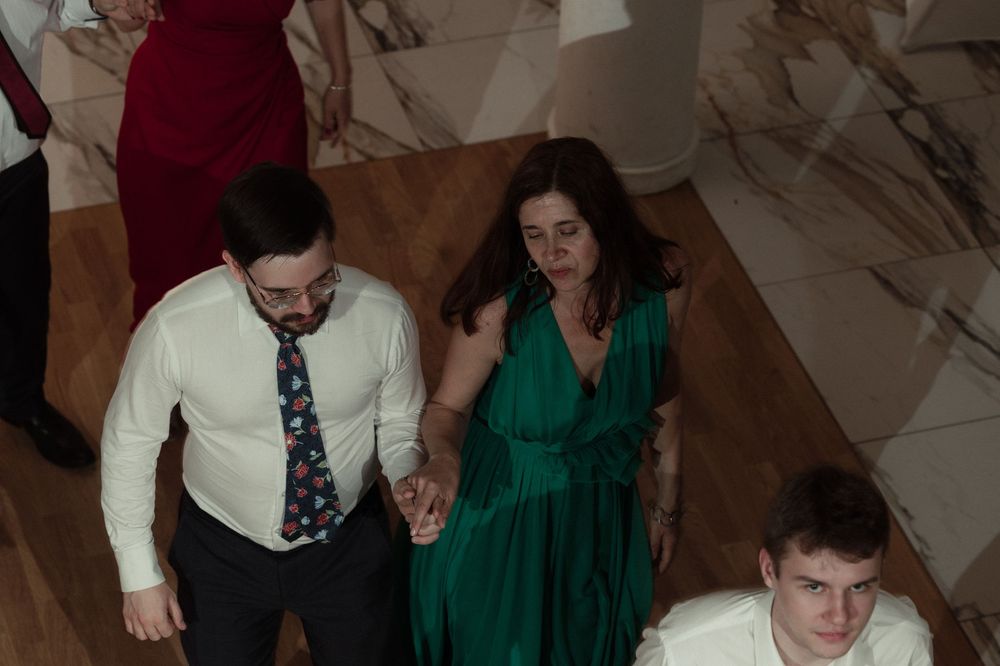 Guests in formal attire dance and socialize at an indoor event with marble walls and brown carpeting.