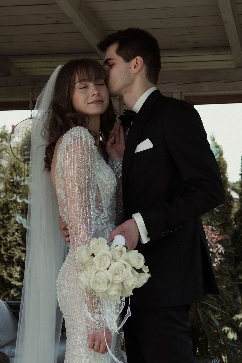 A tender bridal moment captured under a wooden gazebo, featuring a lace long-sleeved gown and classic black tuxedo.