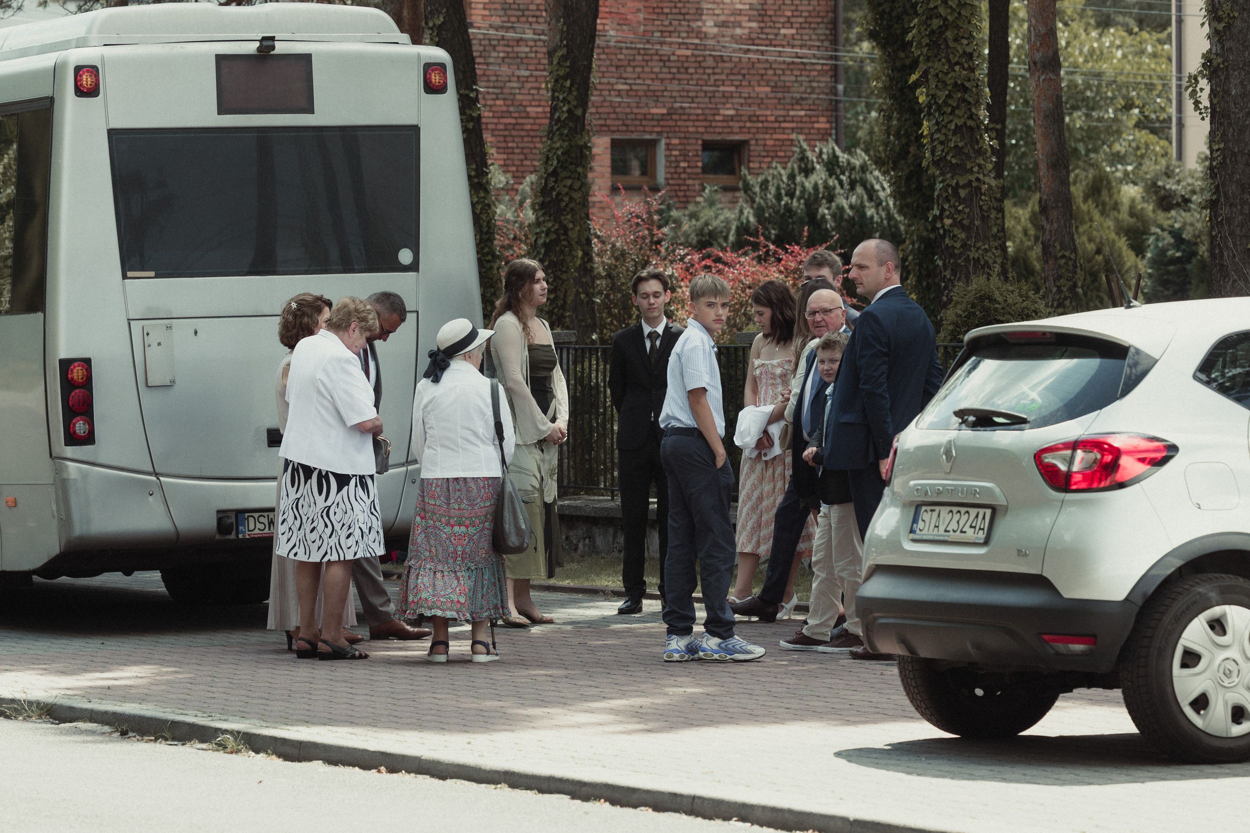 People waiting at a bus stop near parked cars along a brick building in an urban setting.