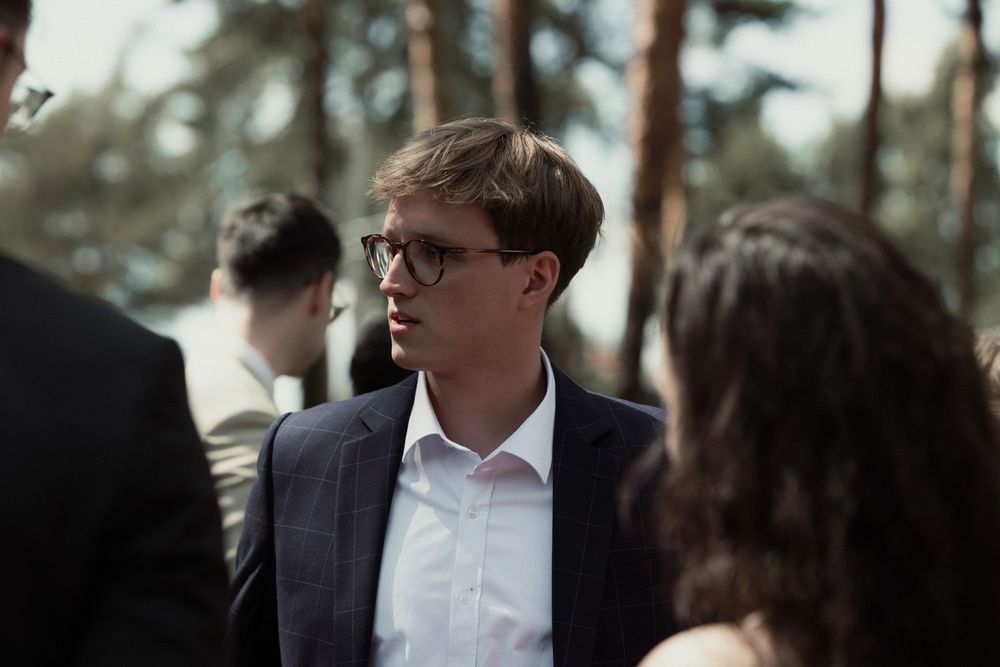People dressed in formal attire gather in an outdoor setting with evergreen trees in the background.
