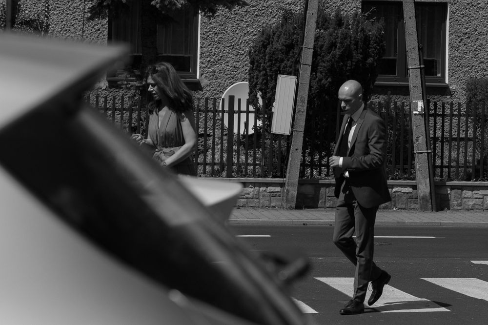 Black and white street scene showing a person getting into a car while another walks past on a sunny day.