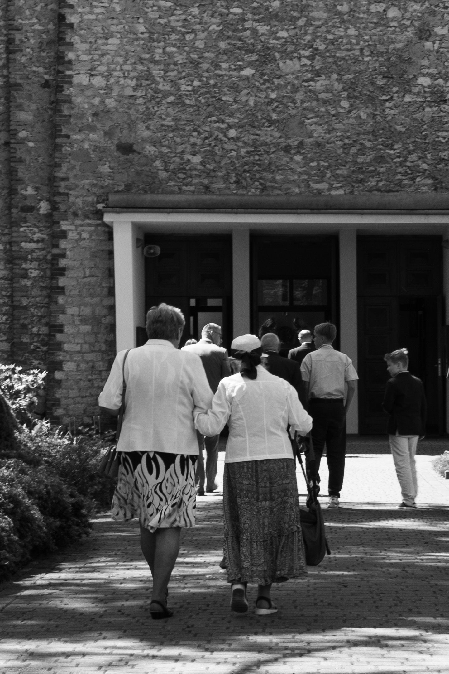Elderly people in casual attire walk together toward a brick building entrance on a sunny day.