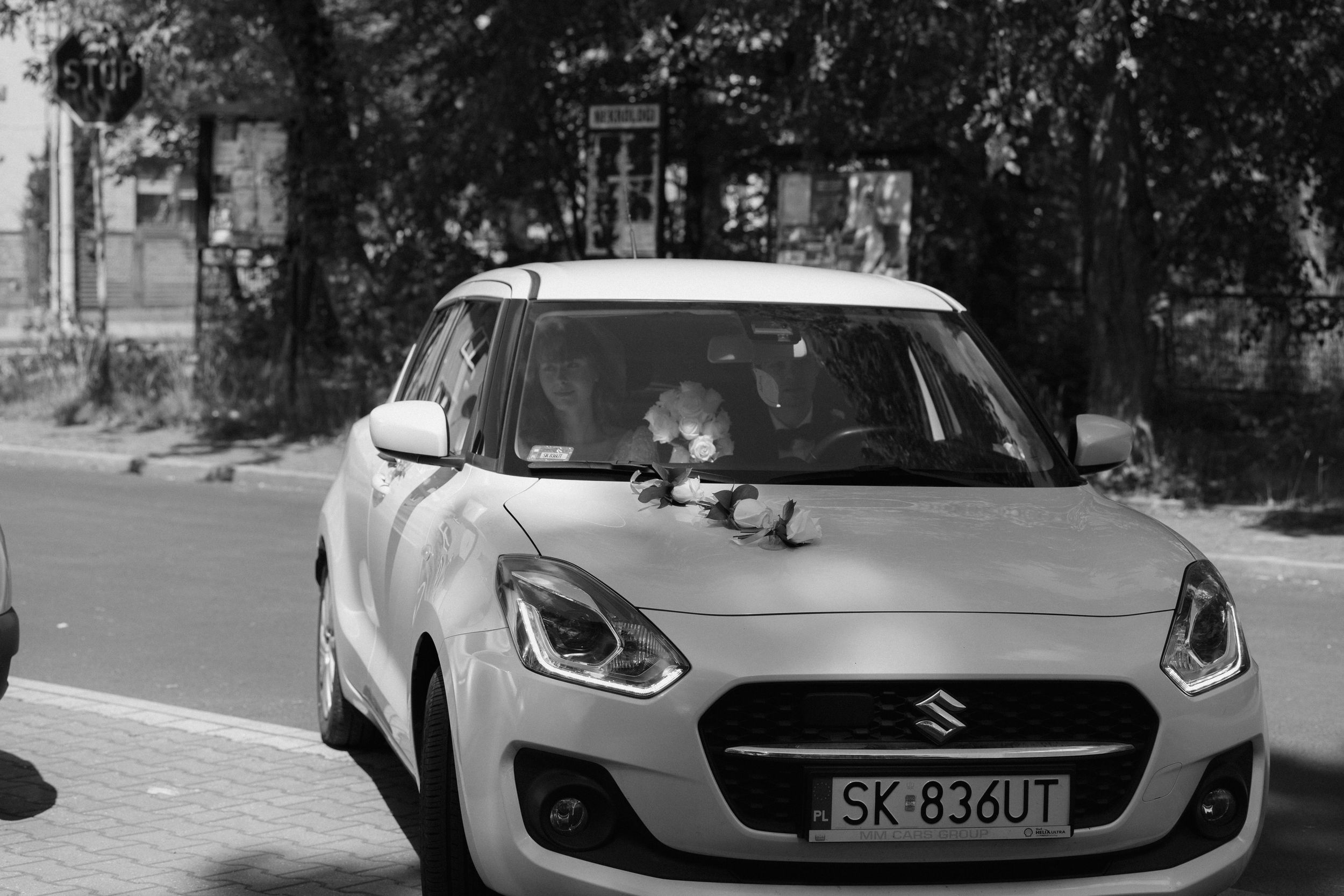 A white Suzuki Swift car parked on a street with trees in the background.