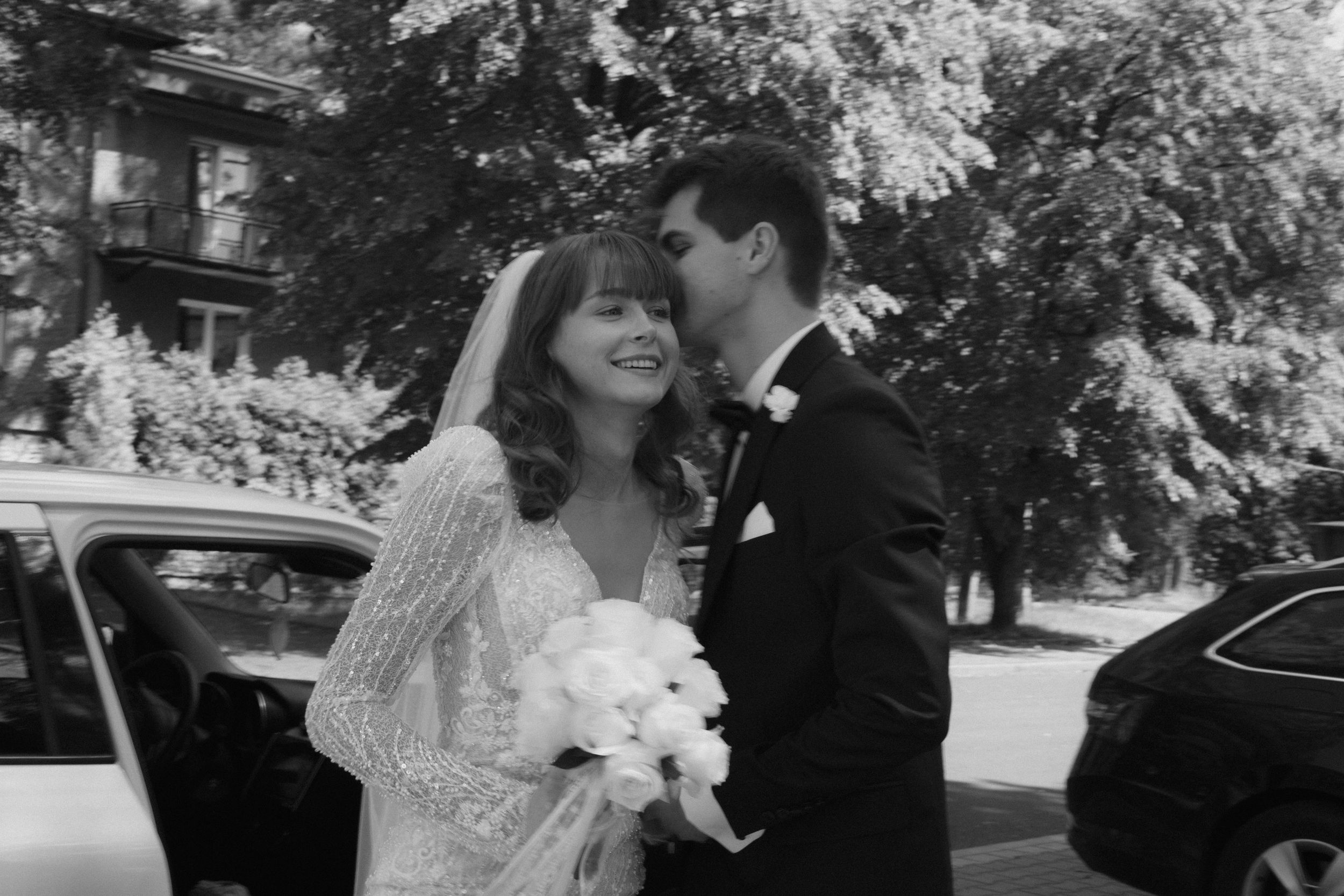 A vintage black and white wedding photo showing a couple by a car holding a white bouquet.