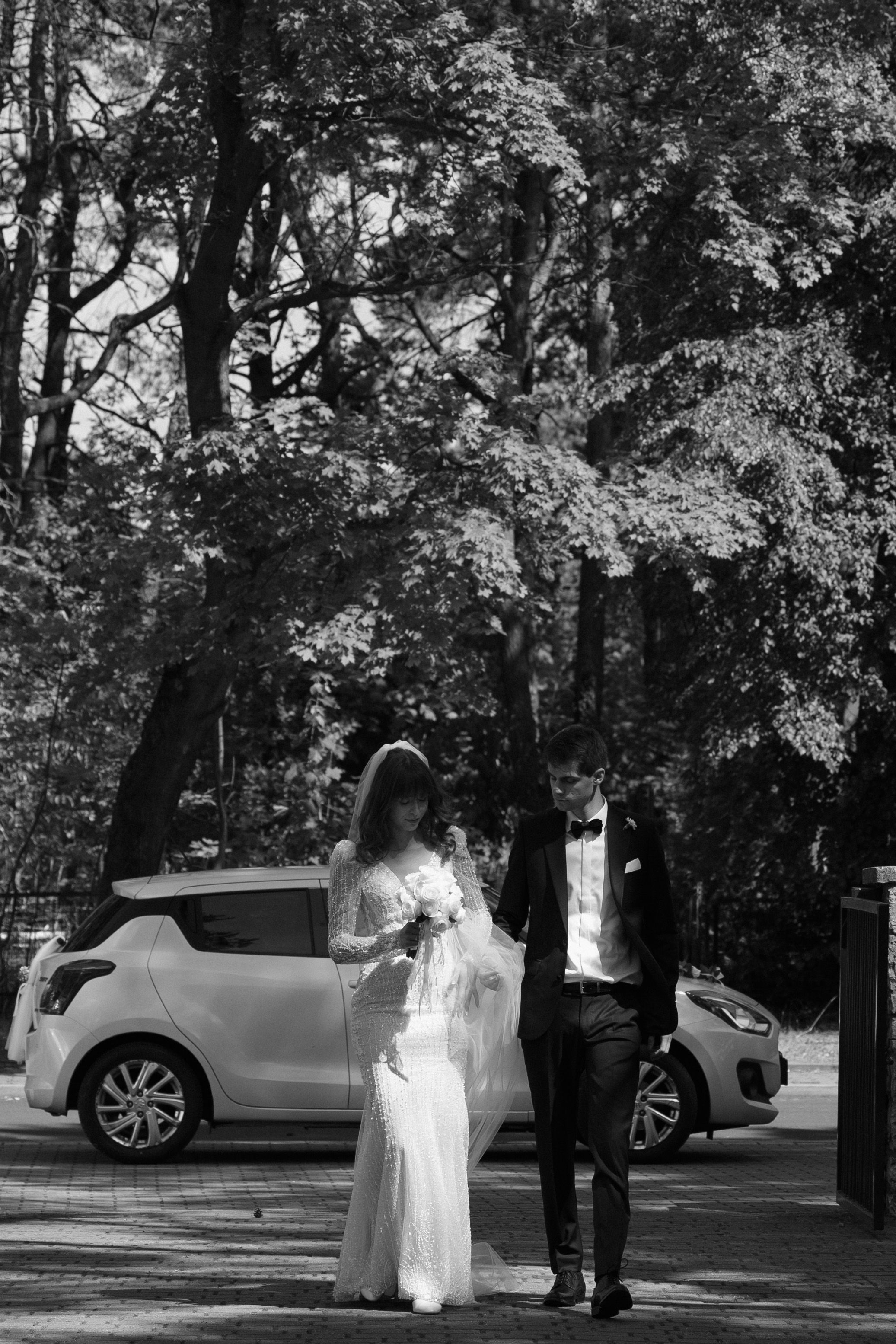 A couple stands outdoors together in formal attire with a white car parked in the background among trees.