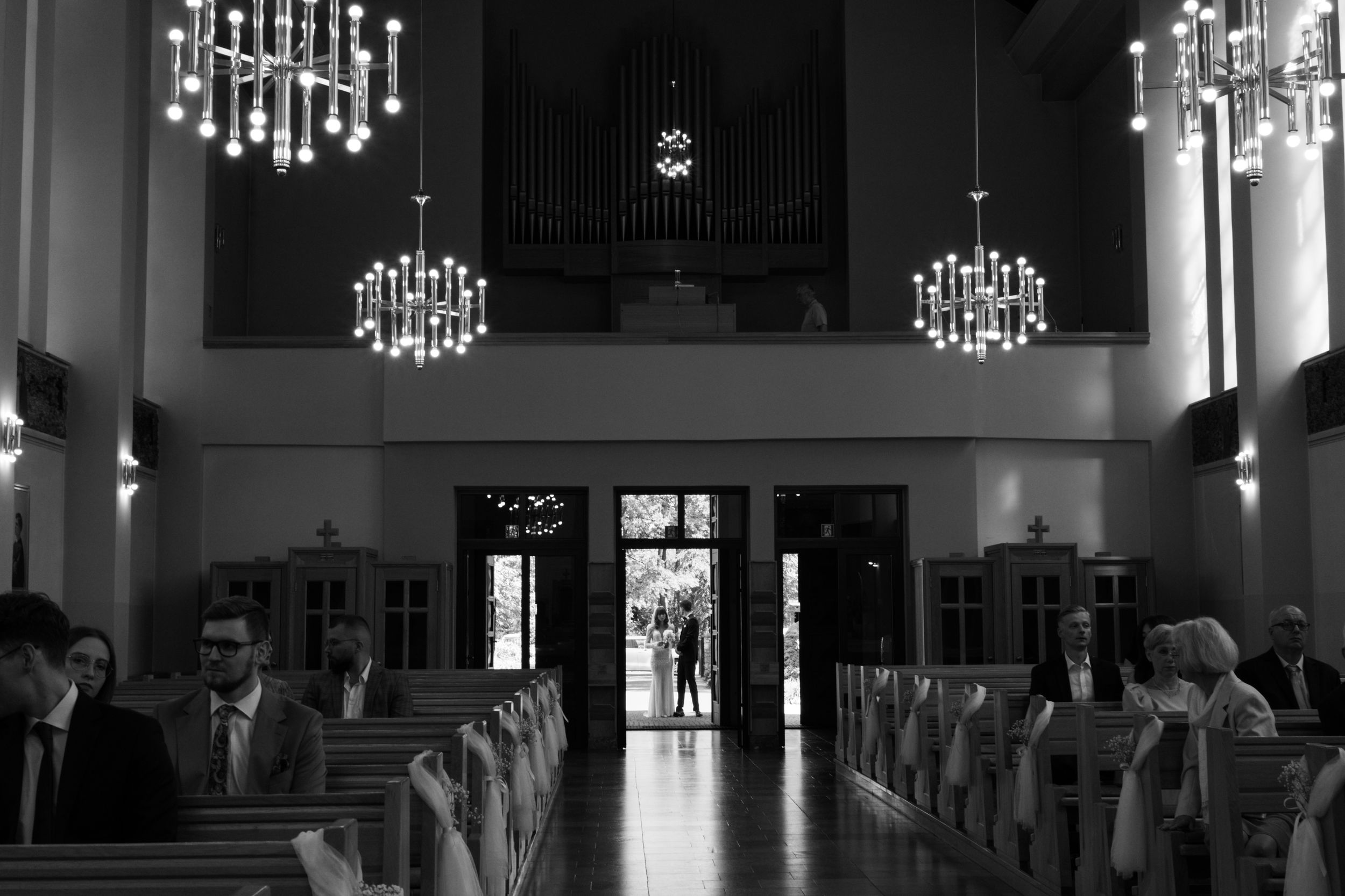 Interior of a church hall with decorative chandeliers and rows of chairs leading to entrance doors.