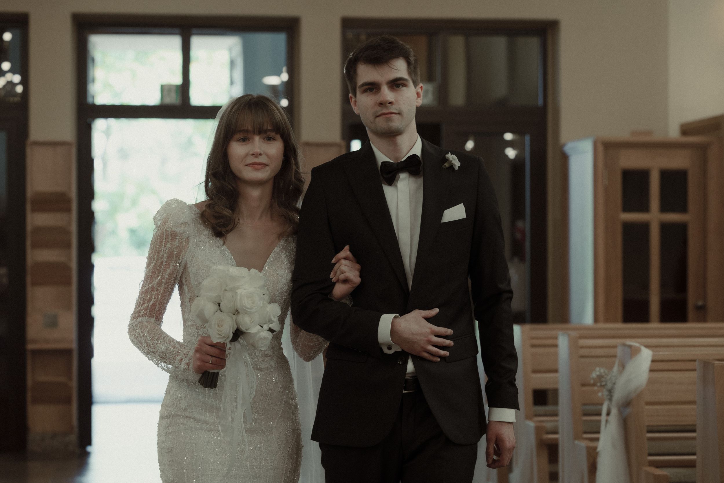 Couple in formal wedding attire stands together in an elegant indoor venue with natural lighting.