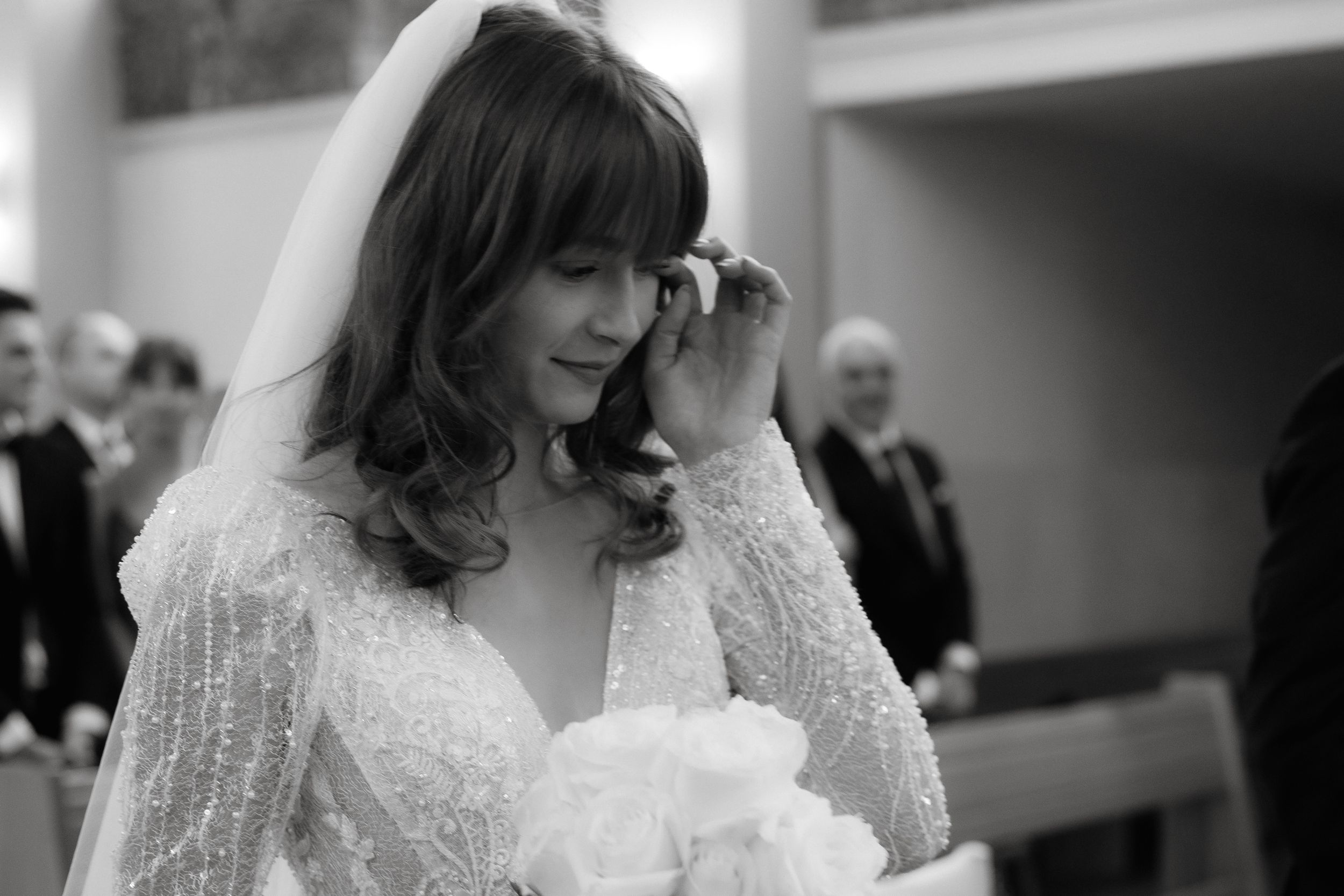 A bride in a white lace wedding dress and veil looks downward while wiping her eye during an emotional ceremony moment.