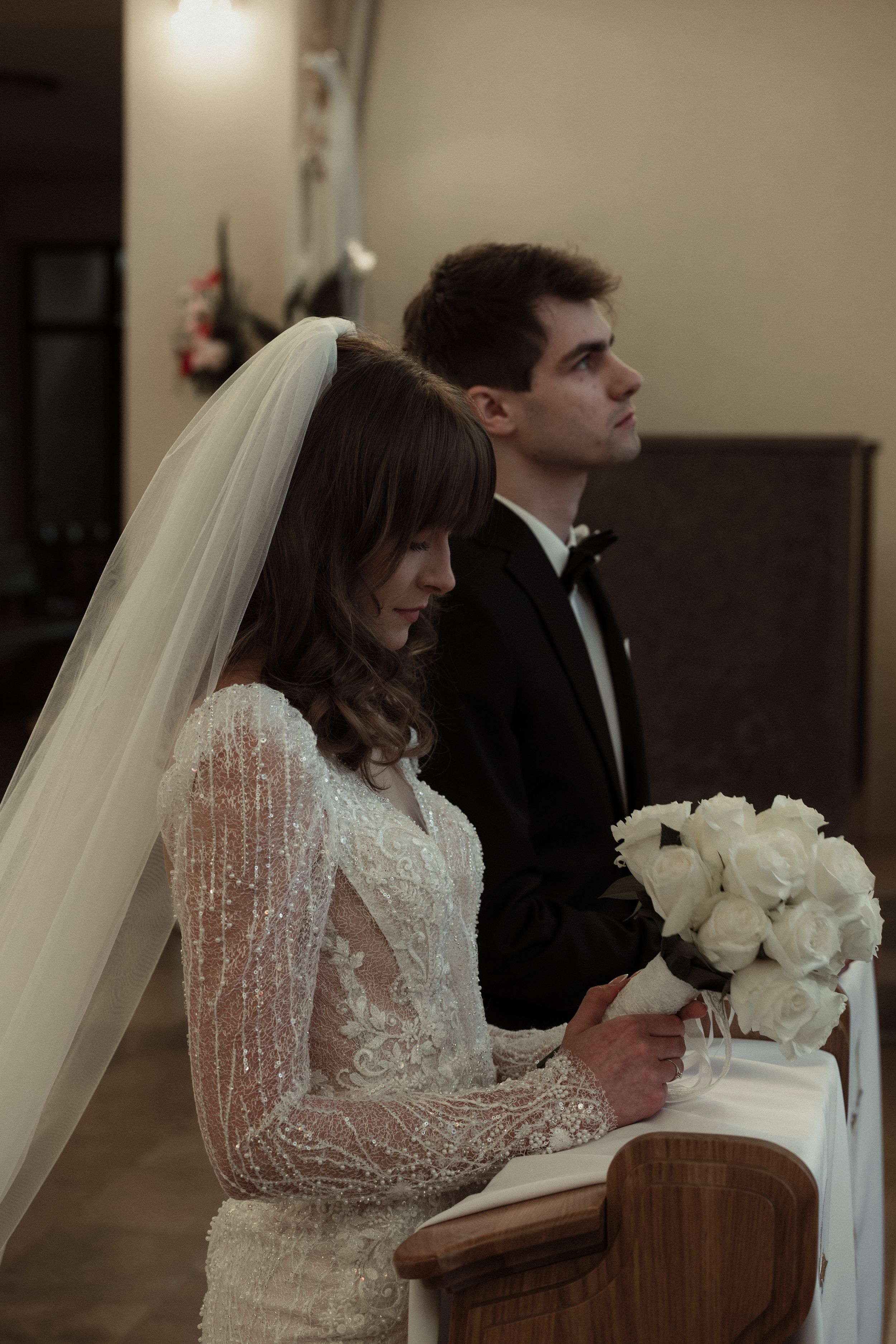 A bride wearing a long-sleeved lace wedding gown and veil holds a white rose bouquet during a church ceremony.