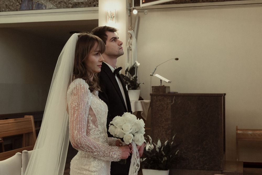 A couple stands during their wedding ceremony in a chapel, with the bride holding a white bouquet.