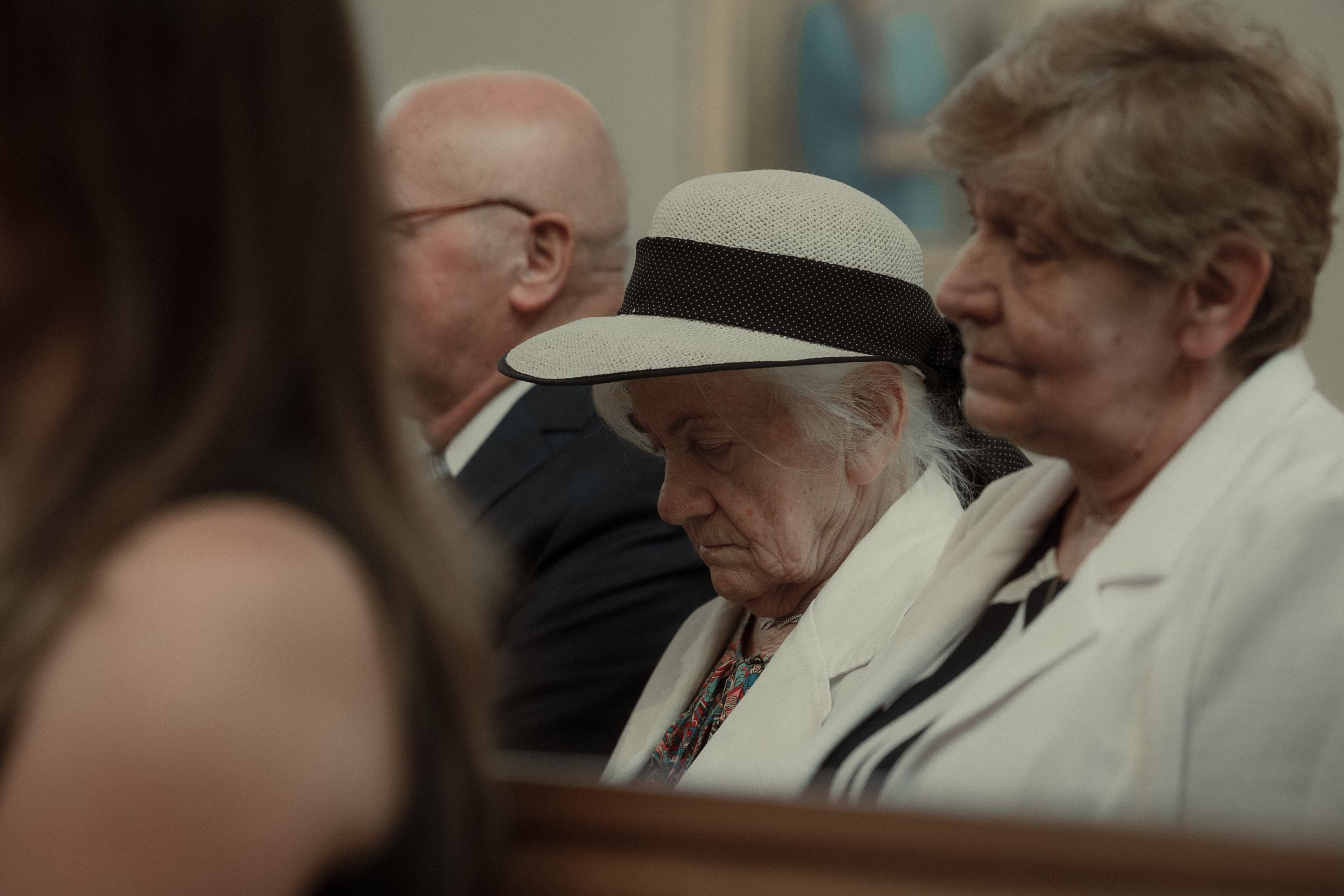 Elderly people sitting together in a church pew wearing light colored clothing and hats.
