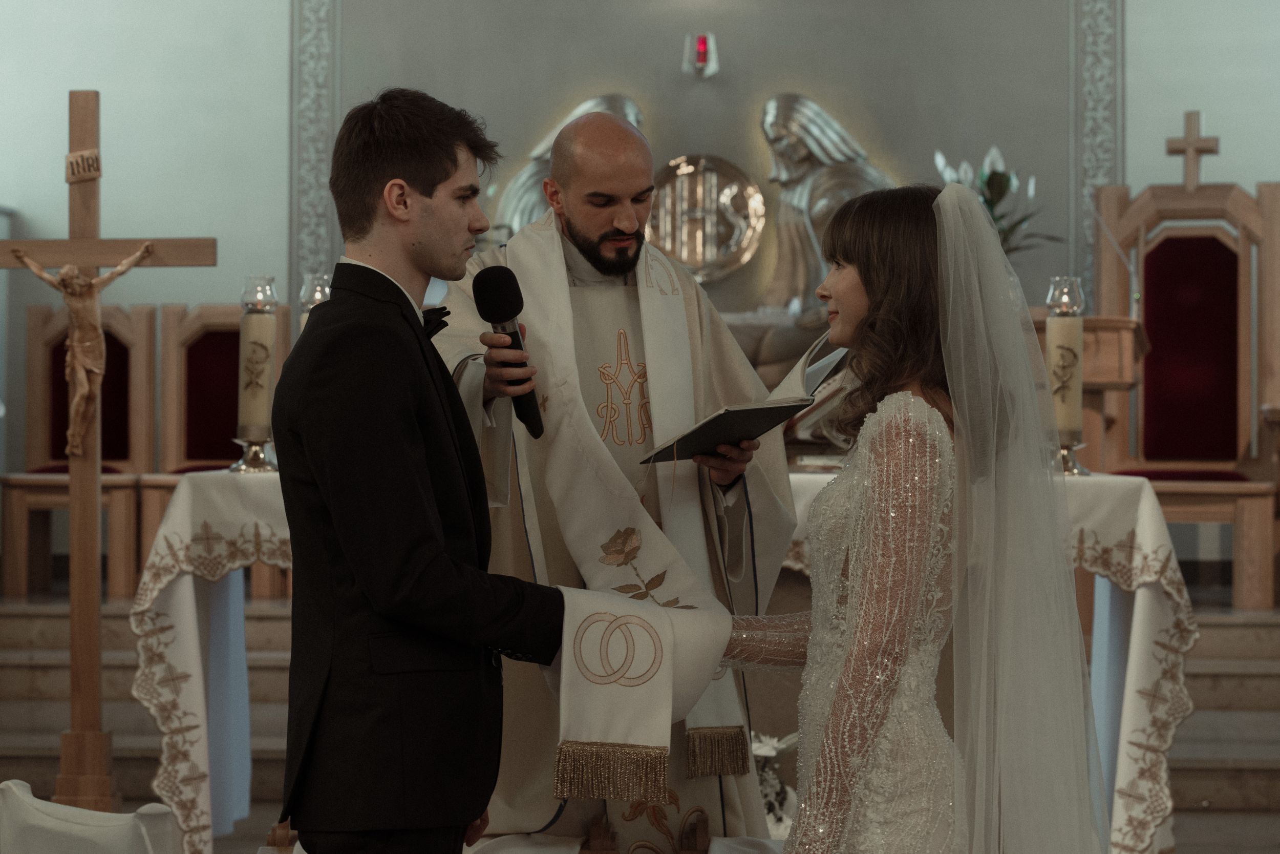 A couple stands at the altar during their wedding ceremony in a church with a religious officiant between them.
