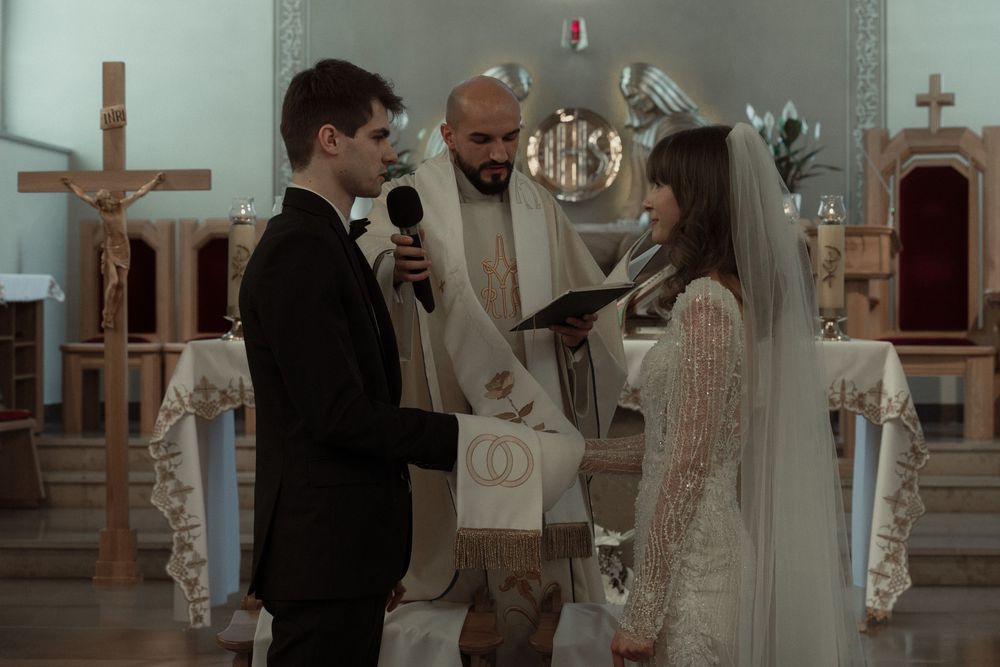 A couple stands at the altar during their wedding ceremony with a priest officiating in a traditional church setting.