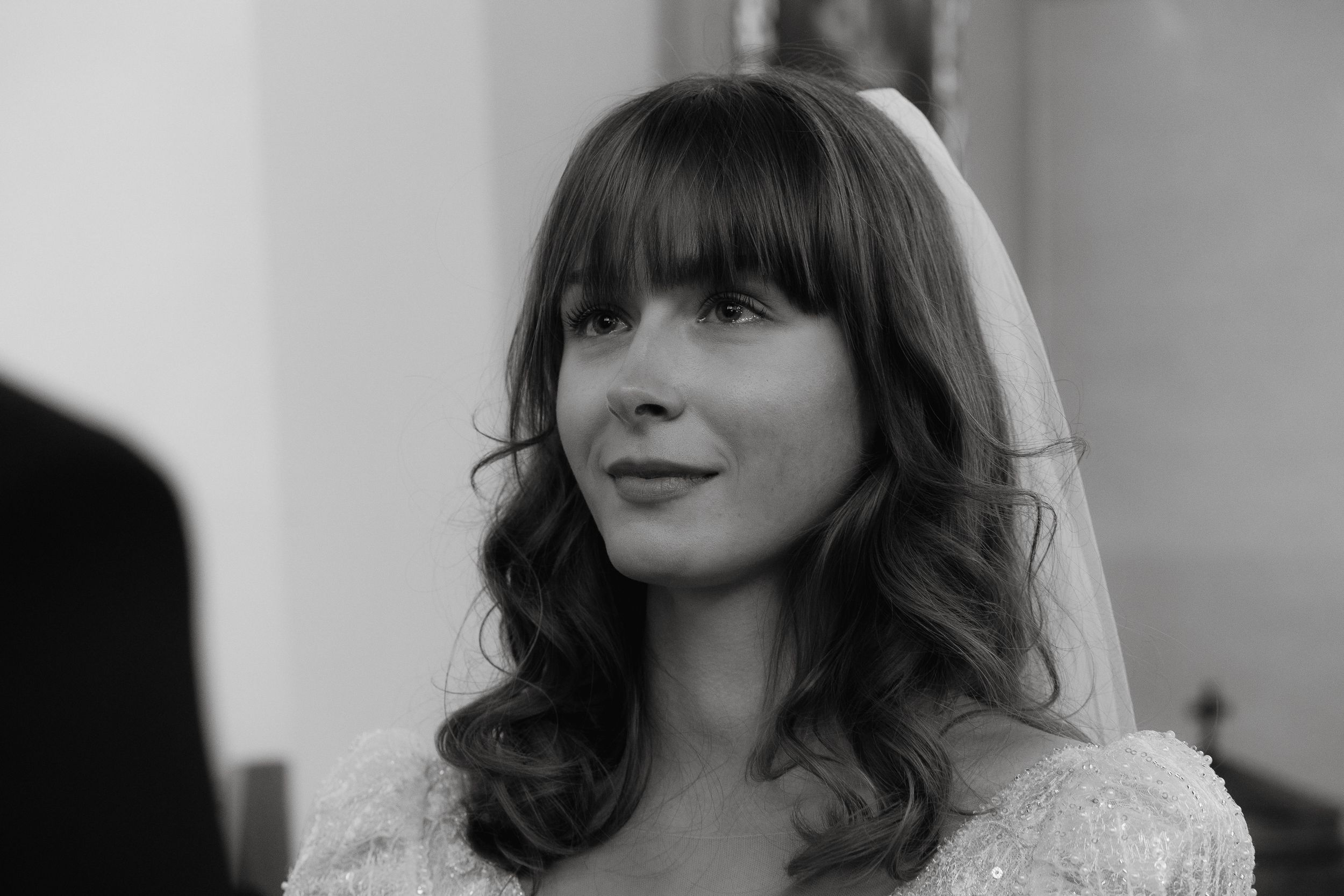 Bride in black and white photograph wearing wedding dress and veil with soft wavy hair and bangs.