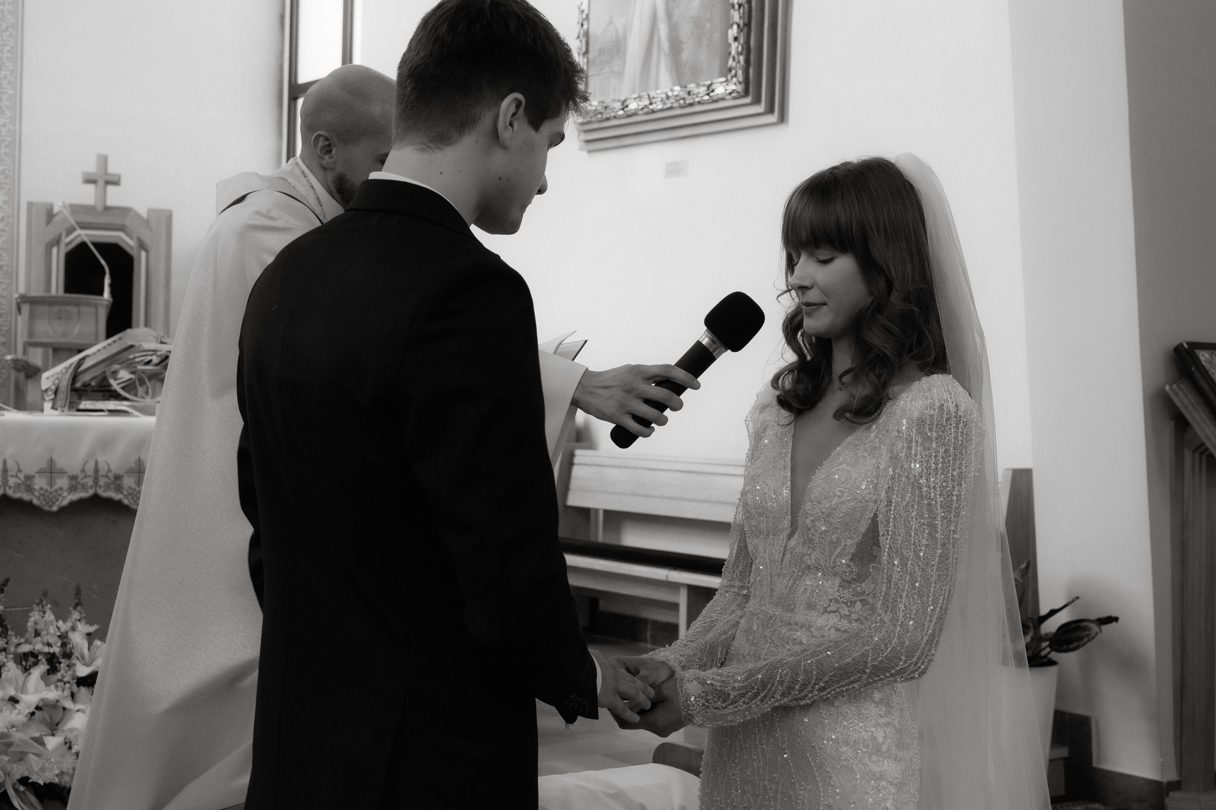 A black and white wedding ceremony photo of two people exchanging vows at the altar in a church.