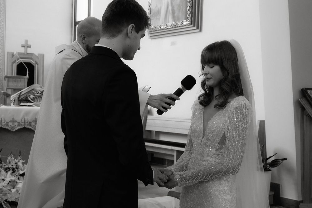 A black and white wedding ceremony photo of two people exchanging vows at the altar in a church.