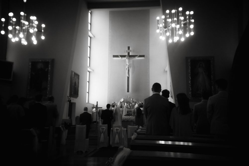 A black and white photograph shows a church interior with a crucifix illuminated by chandeliers during a worship service.