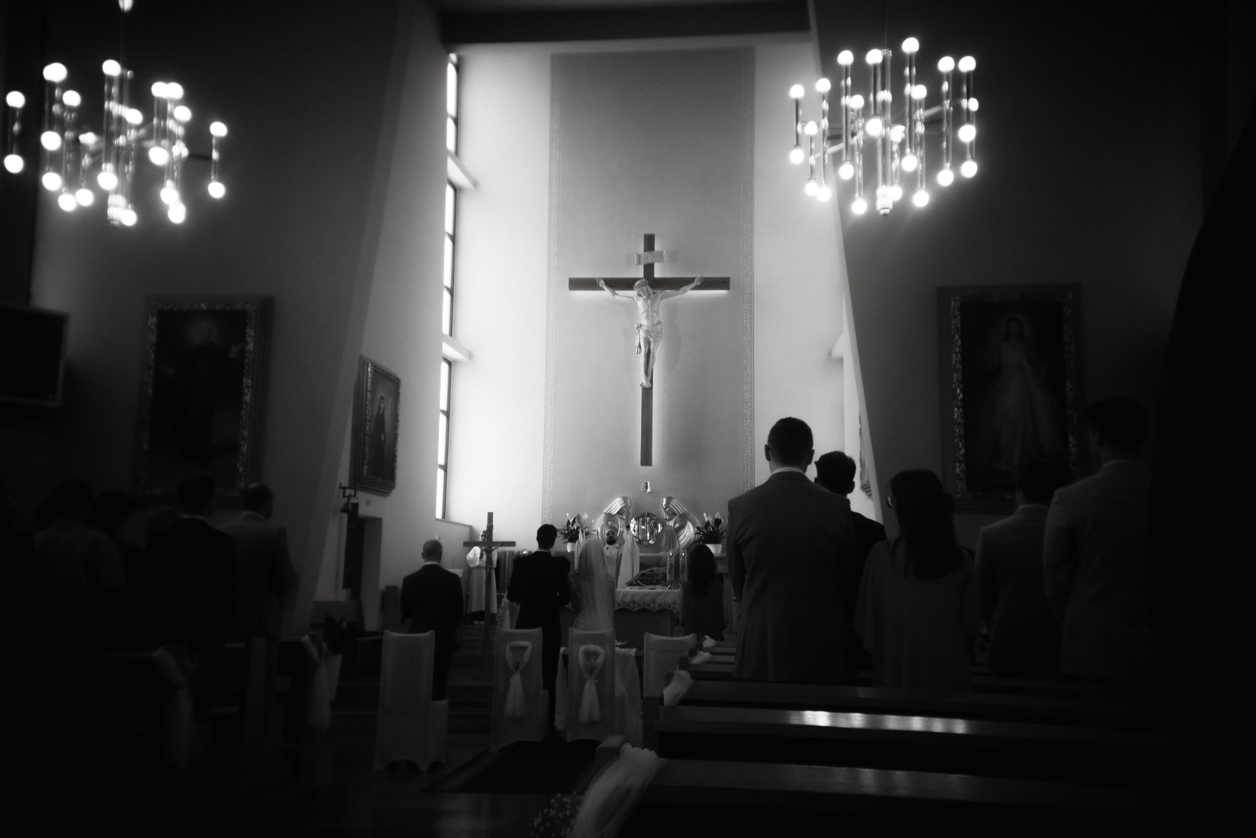A black and white photograph shows a church interior with a crucifix illuminated by chandeliers during a worship service.