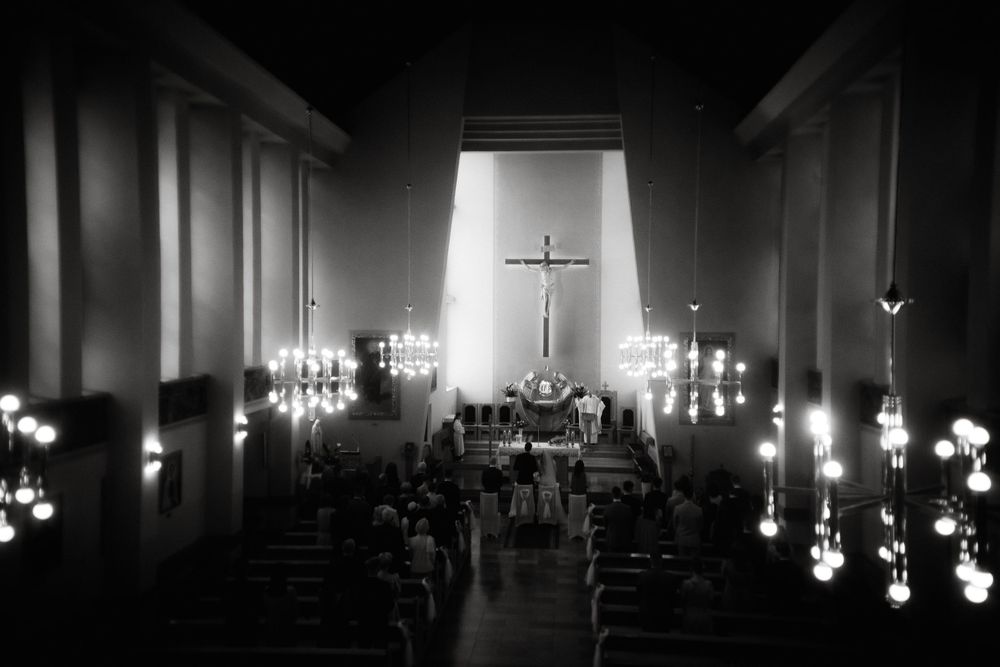 Interior of a dimly lit church with illuminated light fixtures along the walls leading to a glowing cross at the altar.