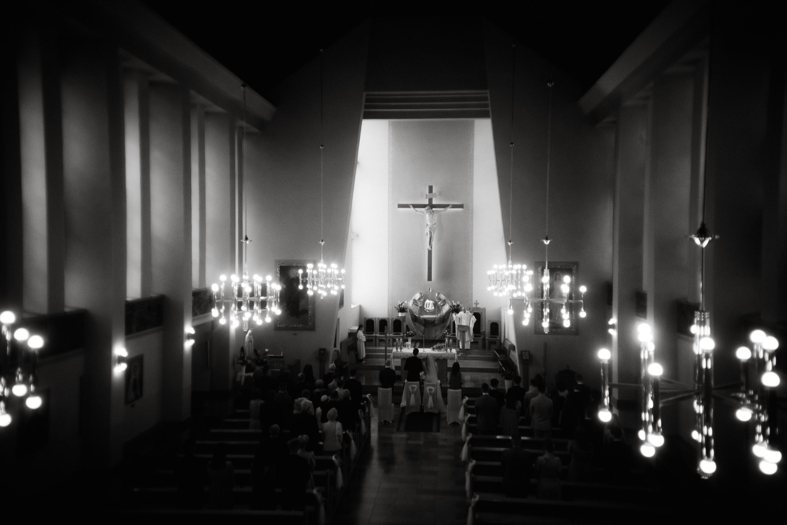 Interior of a dimly lit church with illuminated light fixtures along the walls leading to a glowing cross at the altar.