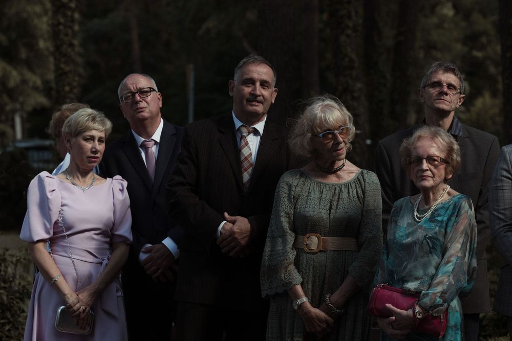 A group of formally dressed guests stand together at an outdoor event wearing suits and formal dresses.
