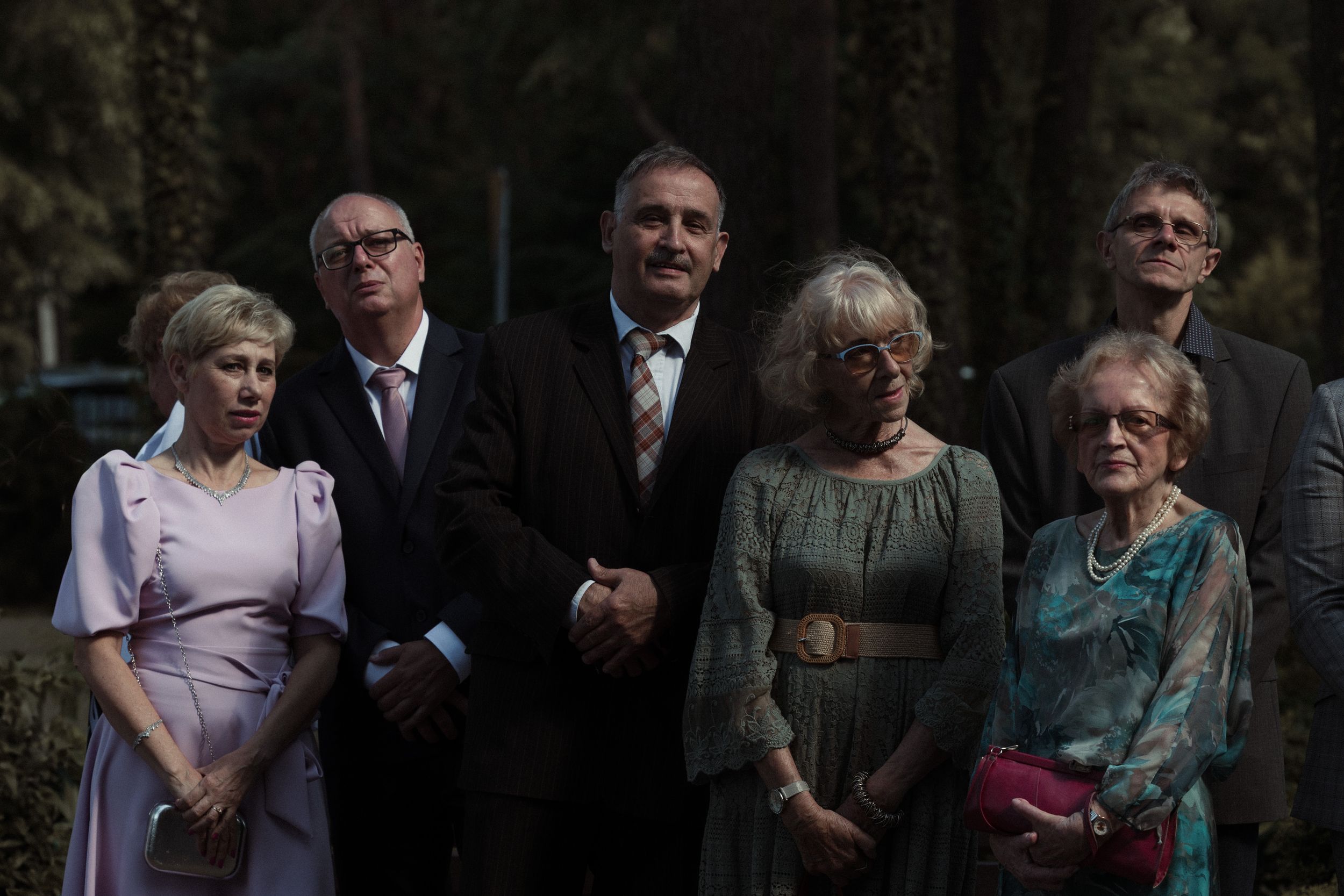 A group of formally dressed guests stand together at an outdoor event wearing suits and formal dresses.