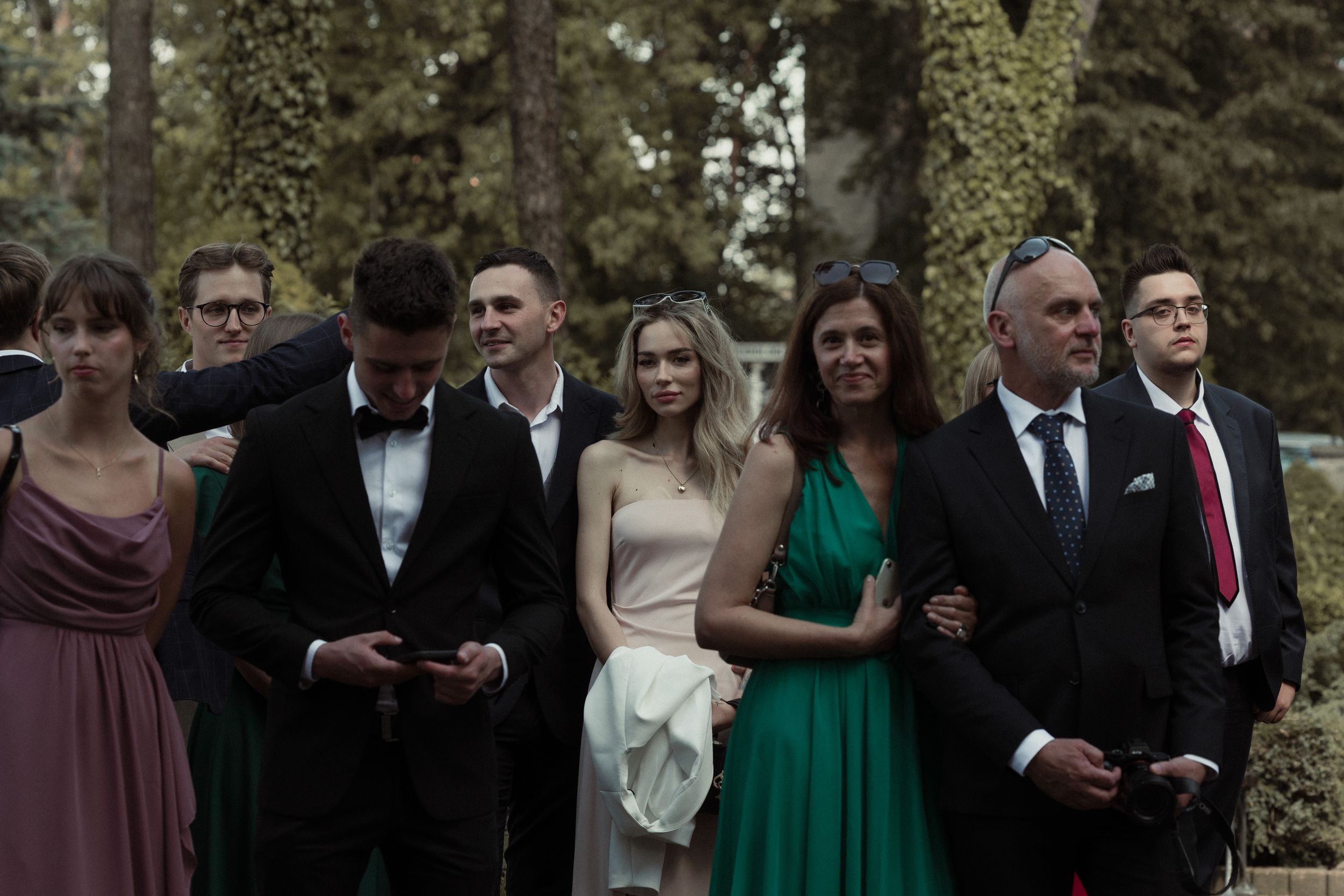 Wedding guests gather outdoors in formal attire including black suits and colorful dresses during a celebration.