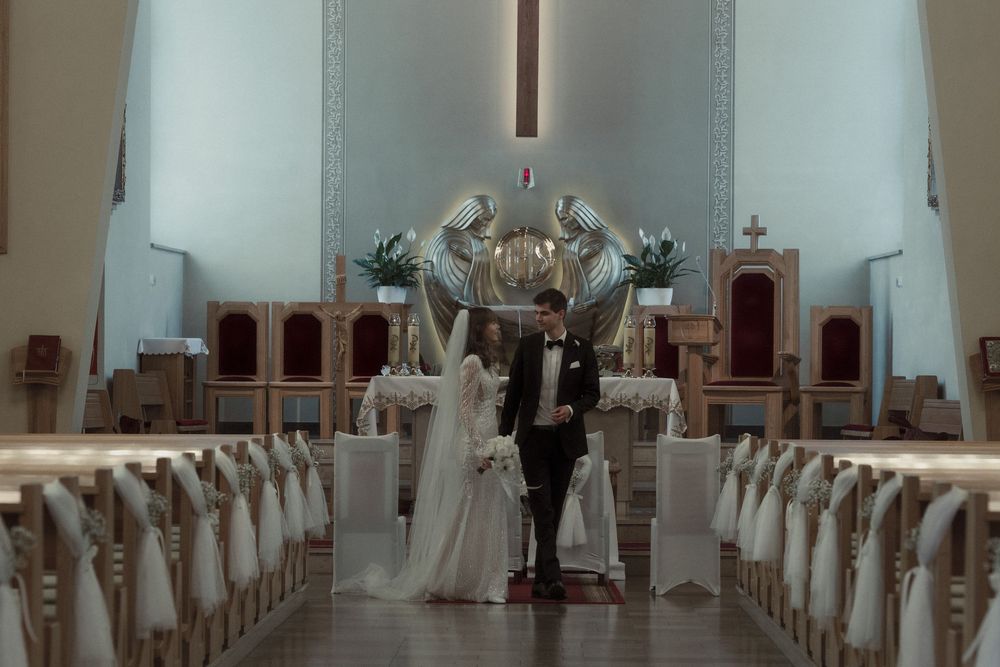 A couple stands together at the front of a bright modern church with white pews and floral arrangements.