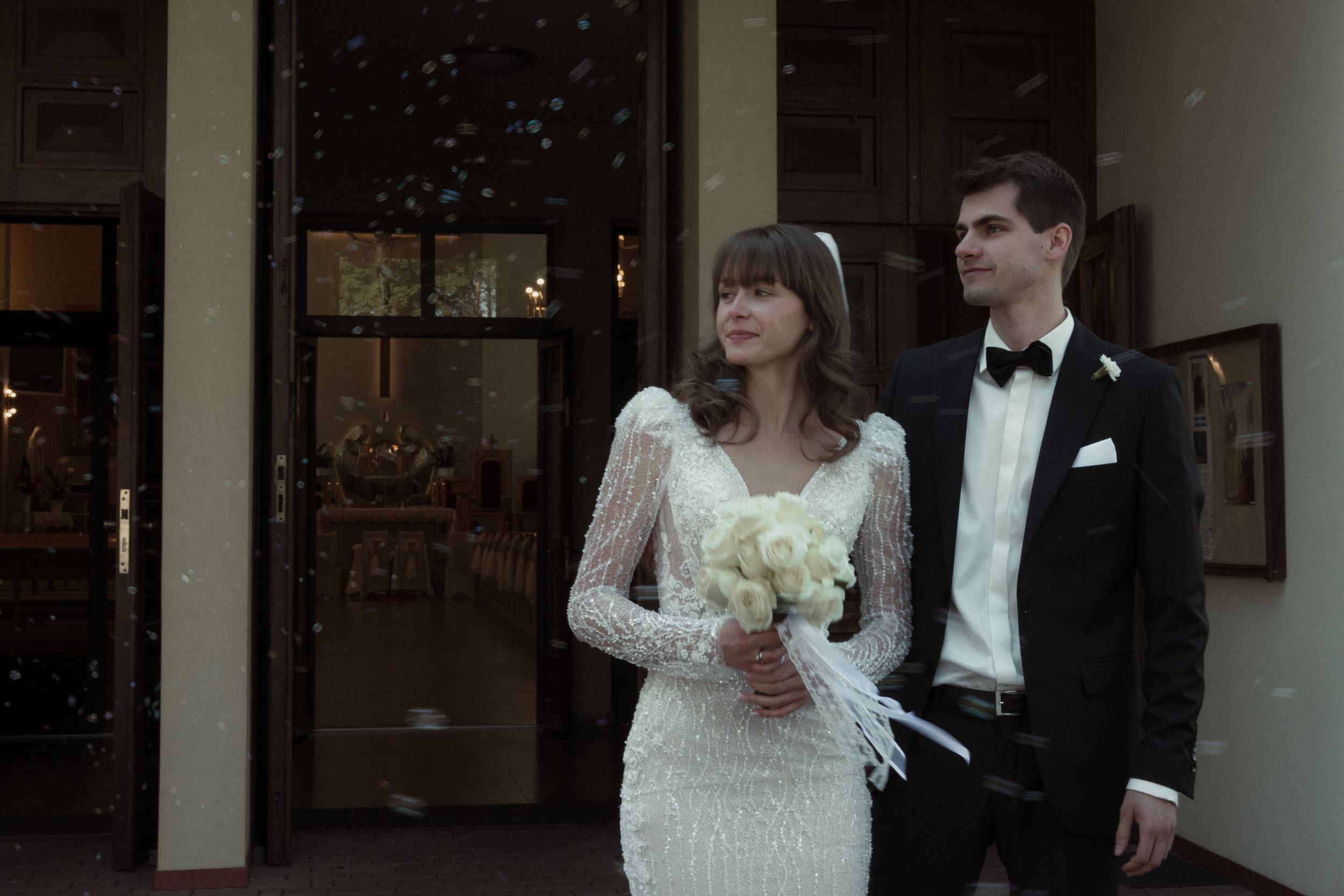 Couple in elegant wedding attire stands together outside a venue at dusk, bride holding a white floral bouquet.
