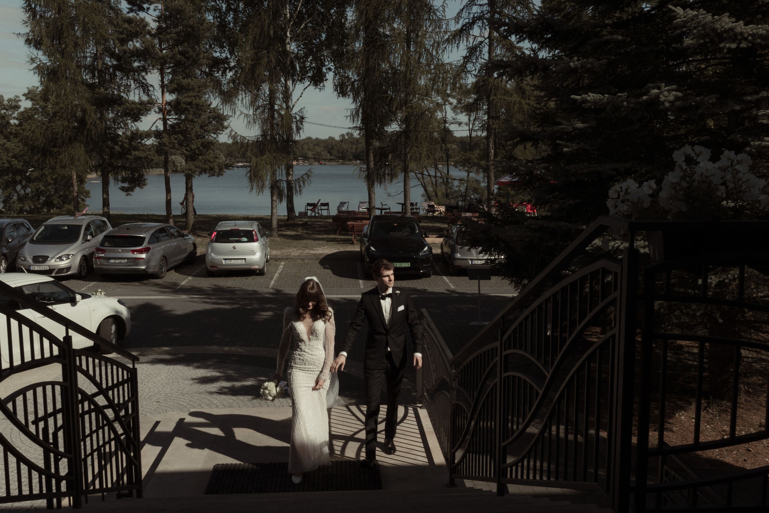 Wedding couple descends outdoor stairs at lakeside venue with parked cars and evergreen trees in background.