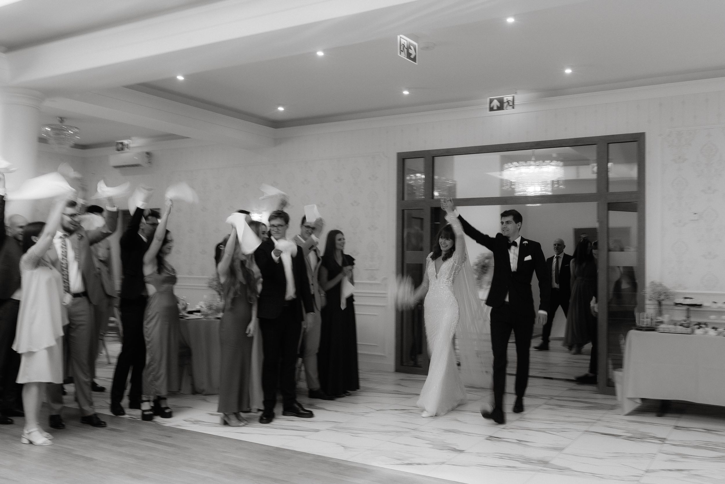 Black and white photo of a festive celebration with people dancing and mingling in a light-filled reception room.