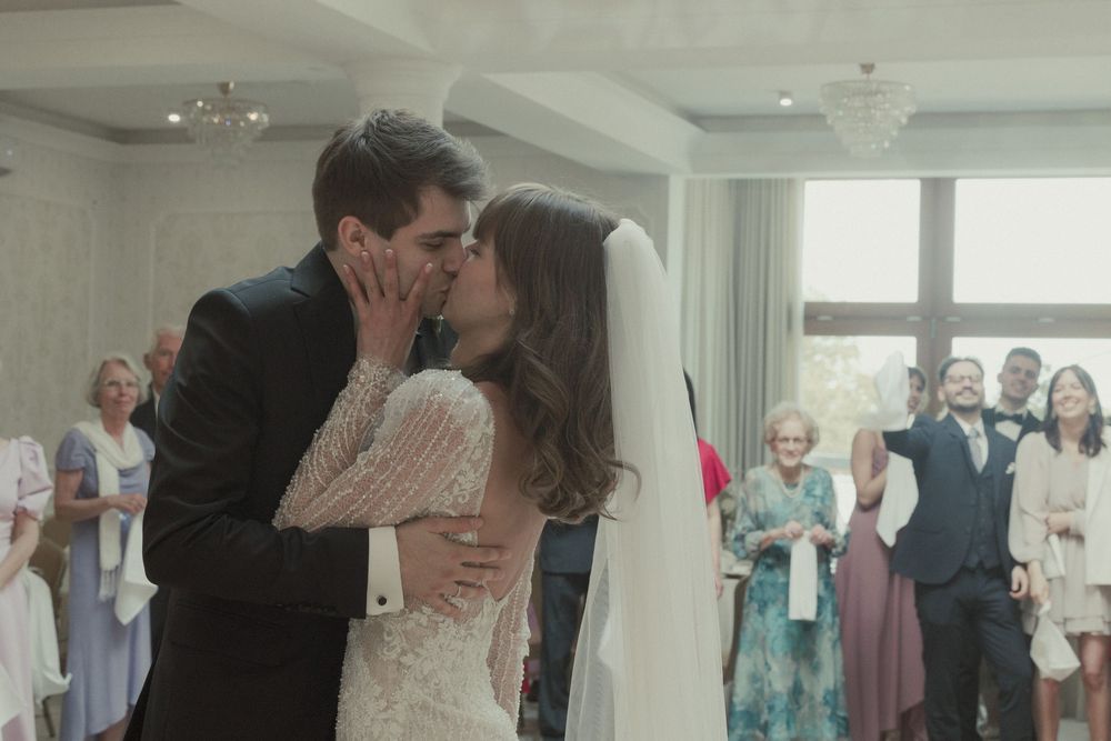 A bride and groom share their first kiss at the altar while wedding guests look on and celebrate in a bright indoor venue.