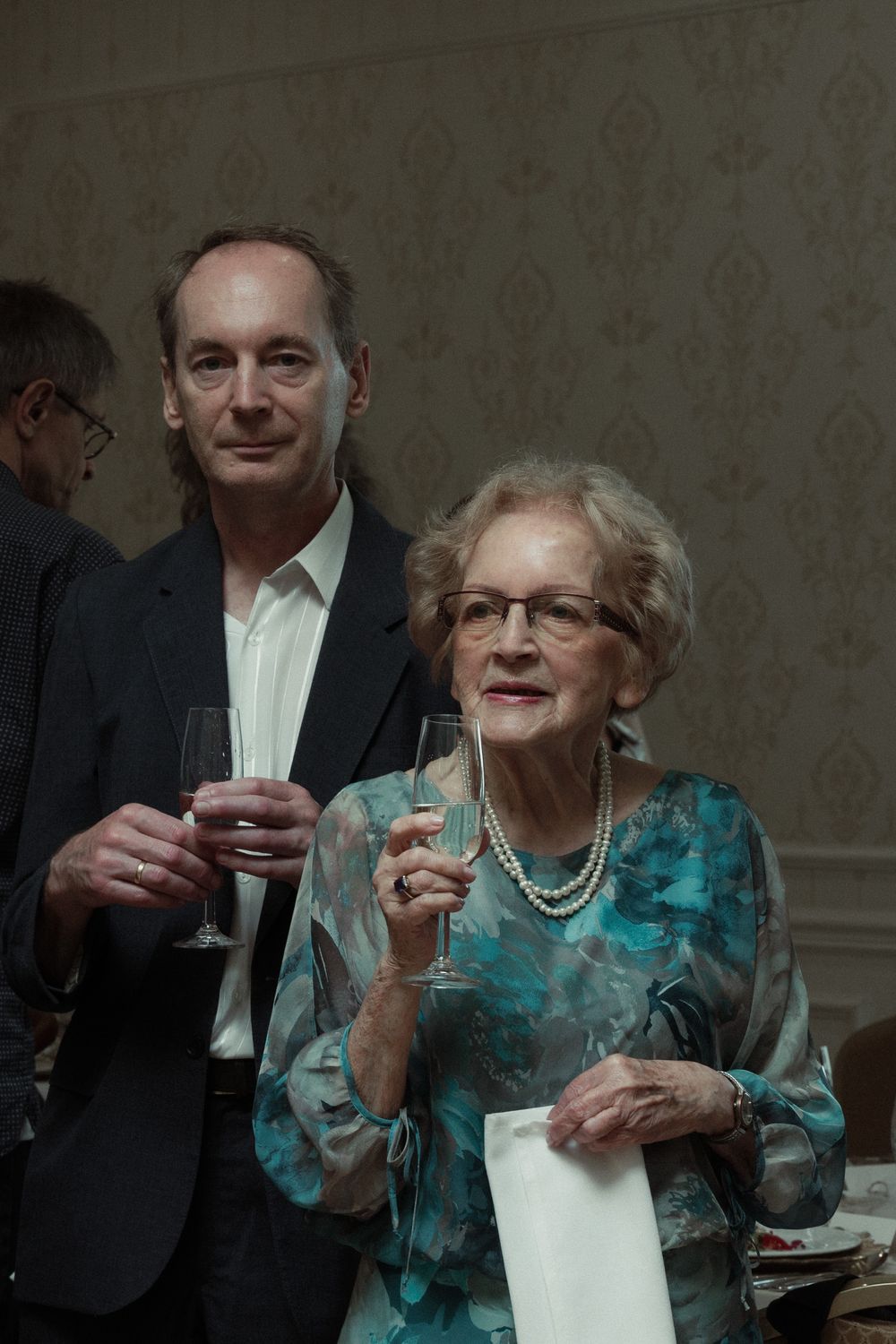 Two people stand together at an elegant event, holding wine glasses and dressed in formal attire against a patterned wall.