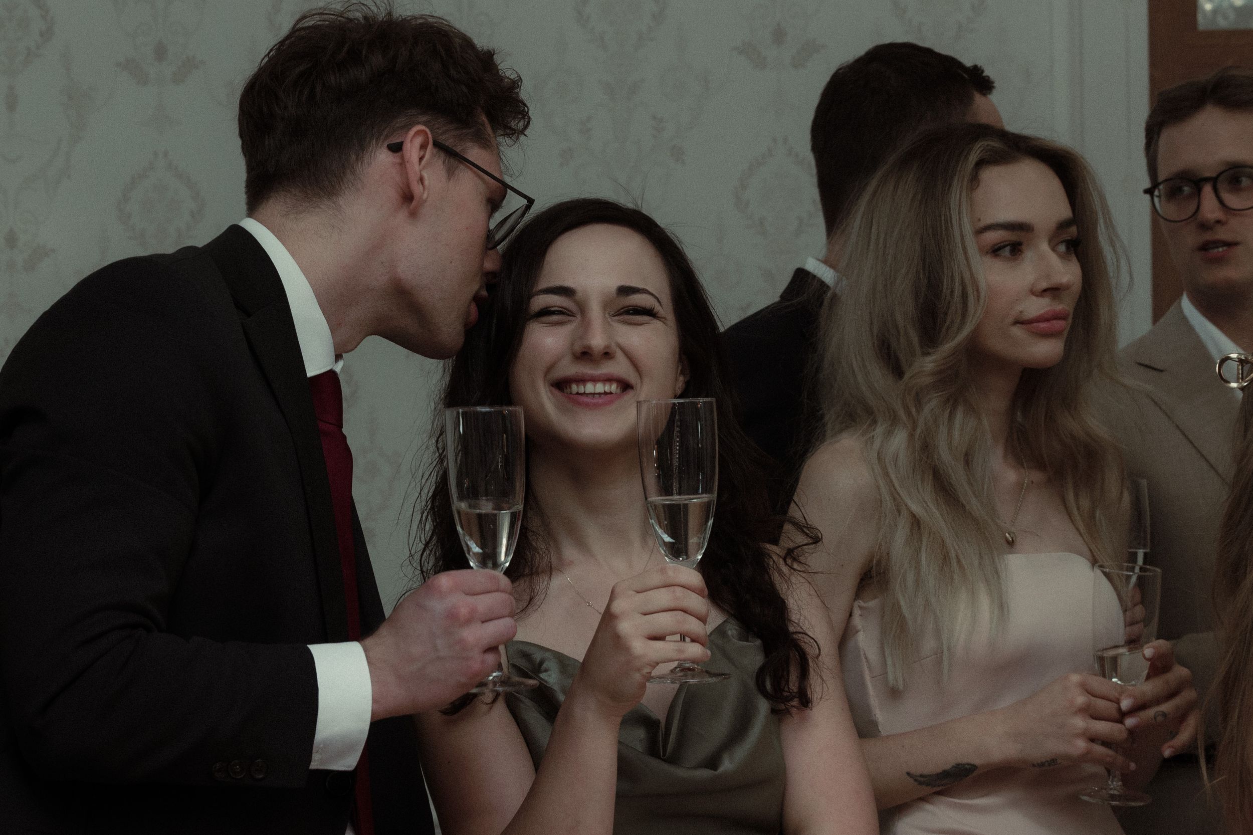 People holding champagne glasses and socializing at an elegant party in dark moody lighting.