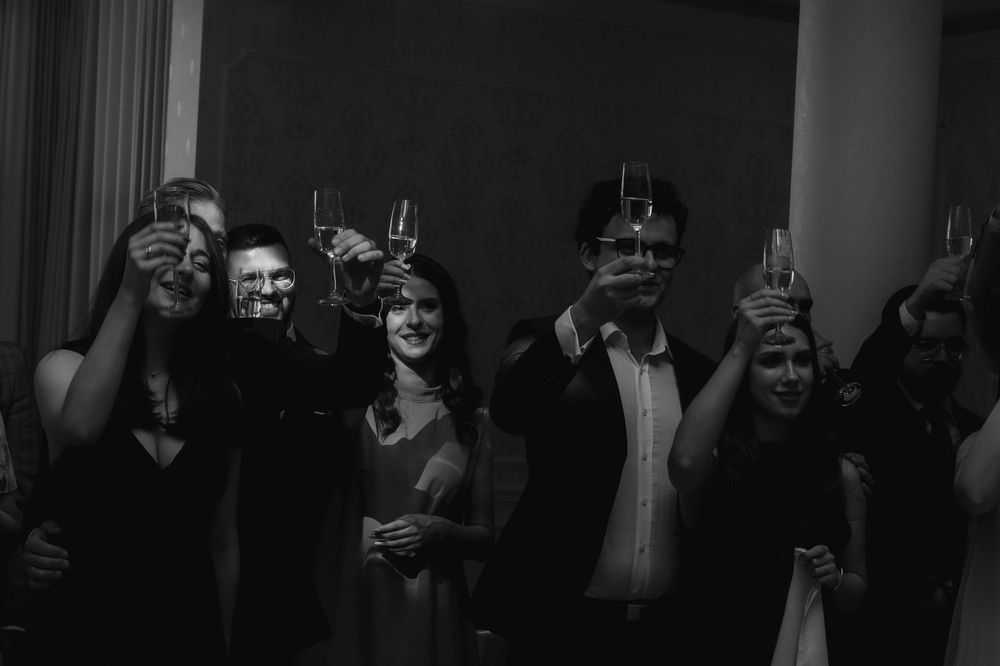 A group of party guests wearing festive masks raise their drinks in a black and white celebratory moment.