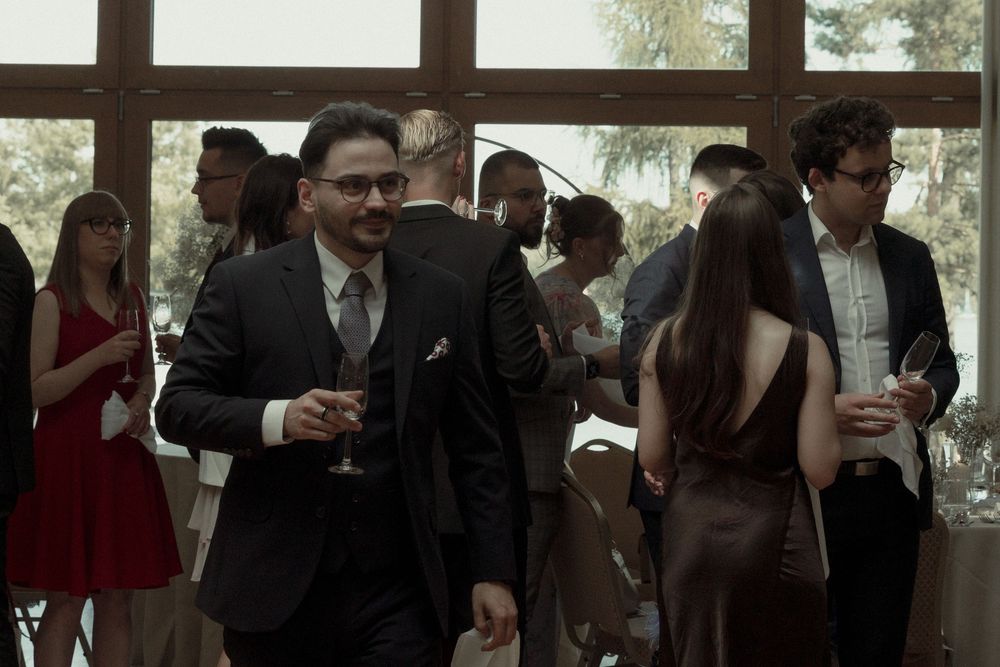 Elegant party guests in formal attire socialize and mingle at an indoor gathering with large windows.