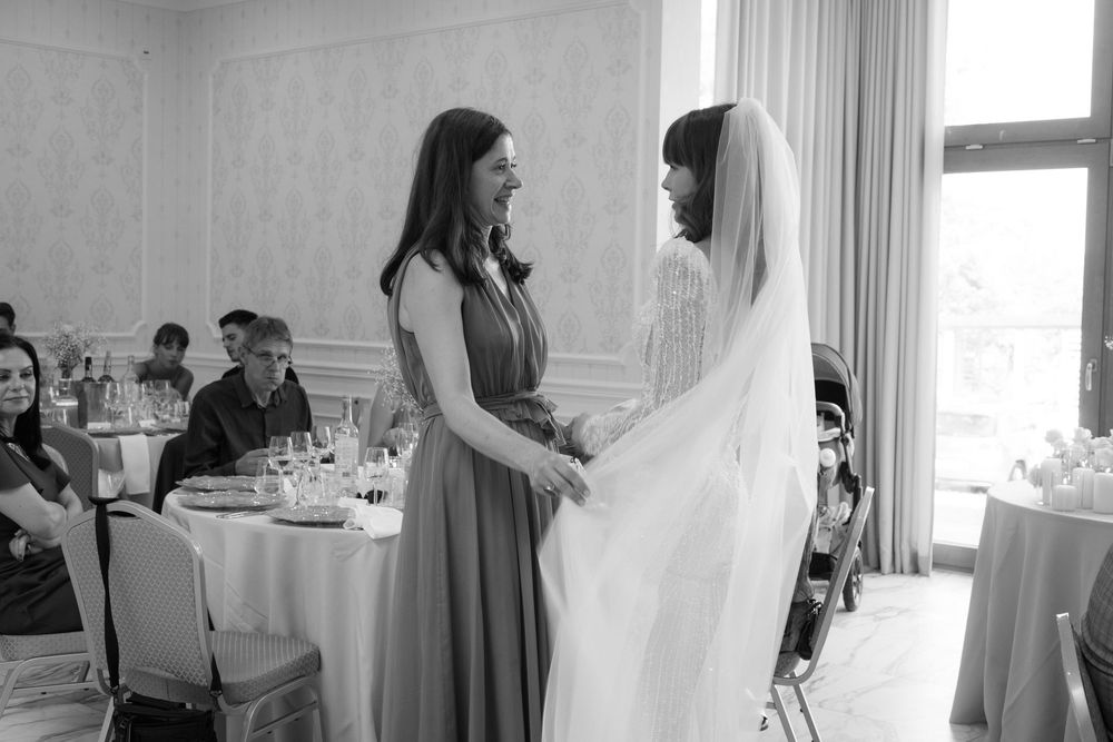 Two women share an intimate moment at a wedding reception, one in a bridesmaid dress and one in a flowing wedding veil.