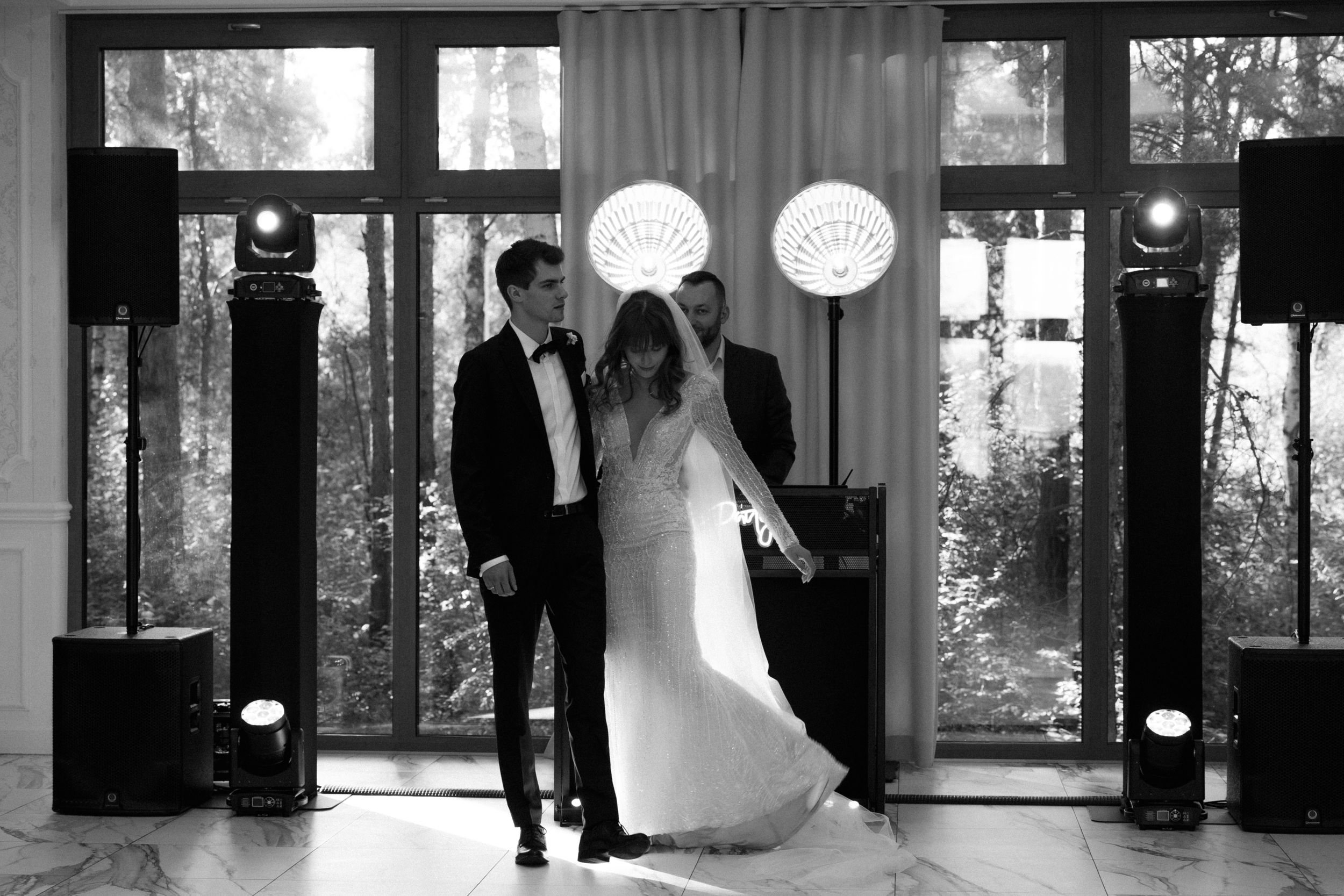 Black and white photo of a couple dancing together at a wedding reception venue with elegant lighting and tall windows.