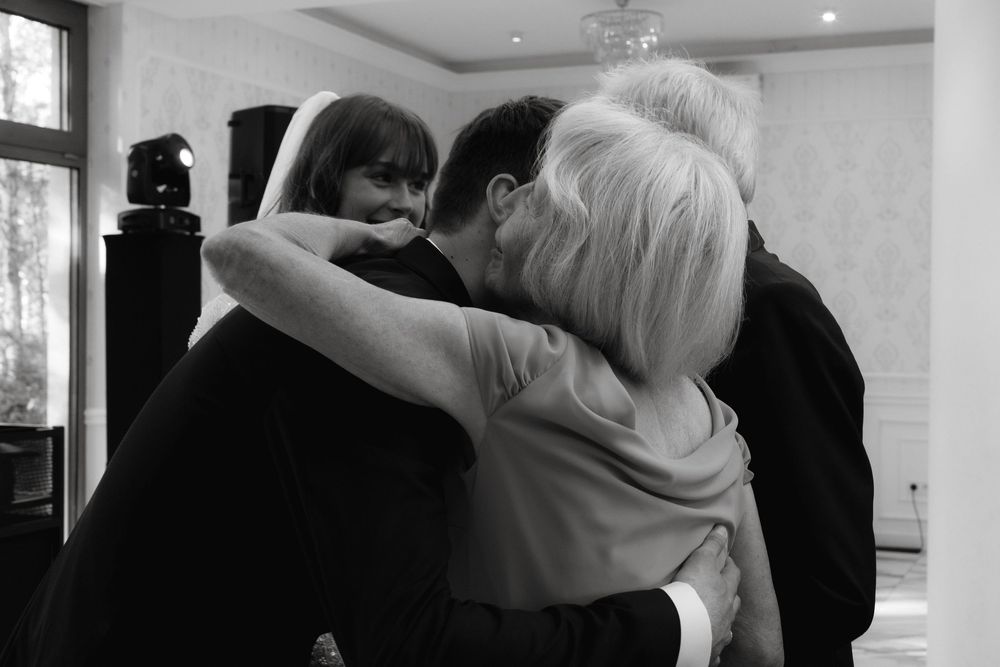 A black and white photograph showing a three person embrace indoors with recessed lighting.