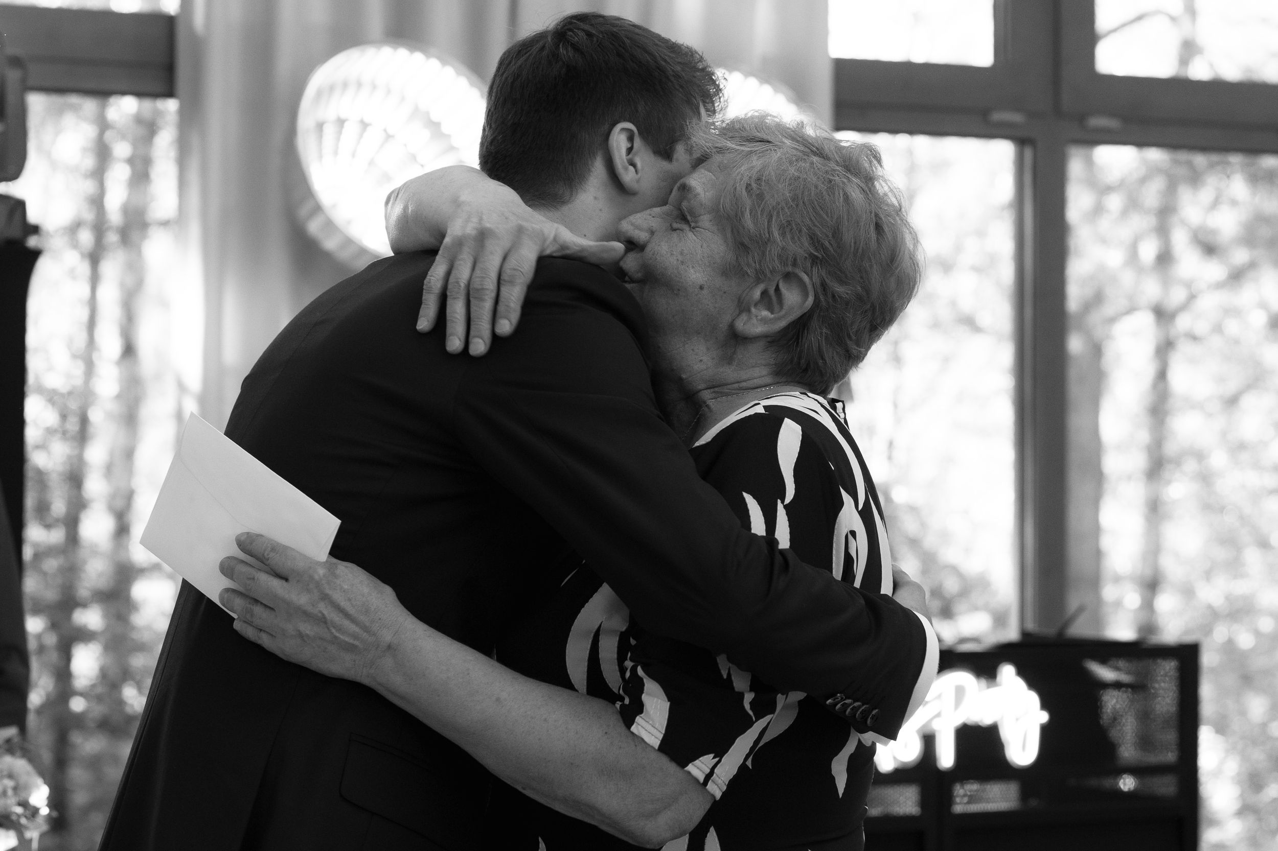 Two elderly individuals embrace in a tender hug near a window, captured in black and white photography.